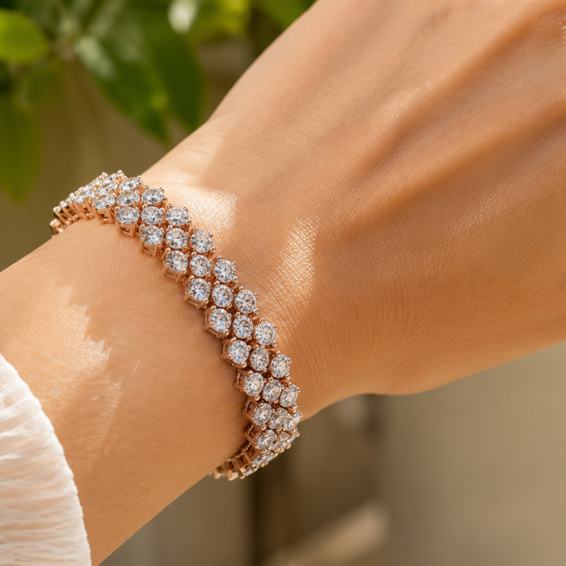 A close-up of a rose gold double-strand bracelet with pavé-set clear stones worn on a wrist, with a soft-focus background of green leaves and neutral tones.