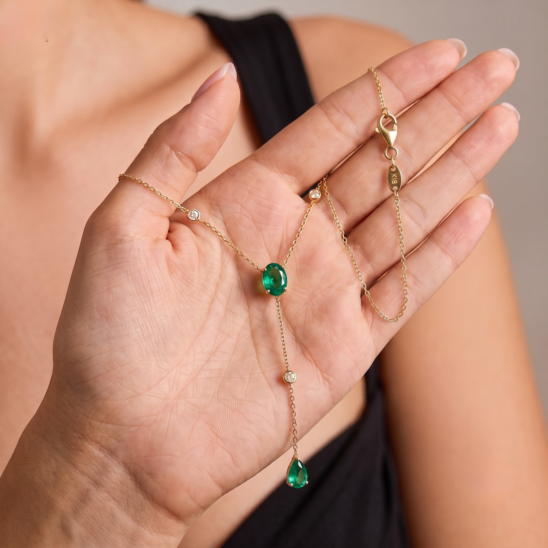 A delicate gold chain necklace with a central round green emerald and two smaller green teardrop drops, held in a person's hand. The necklace also features tiny clear accent stones on the chain.
