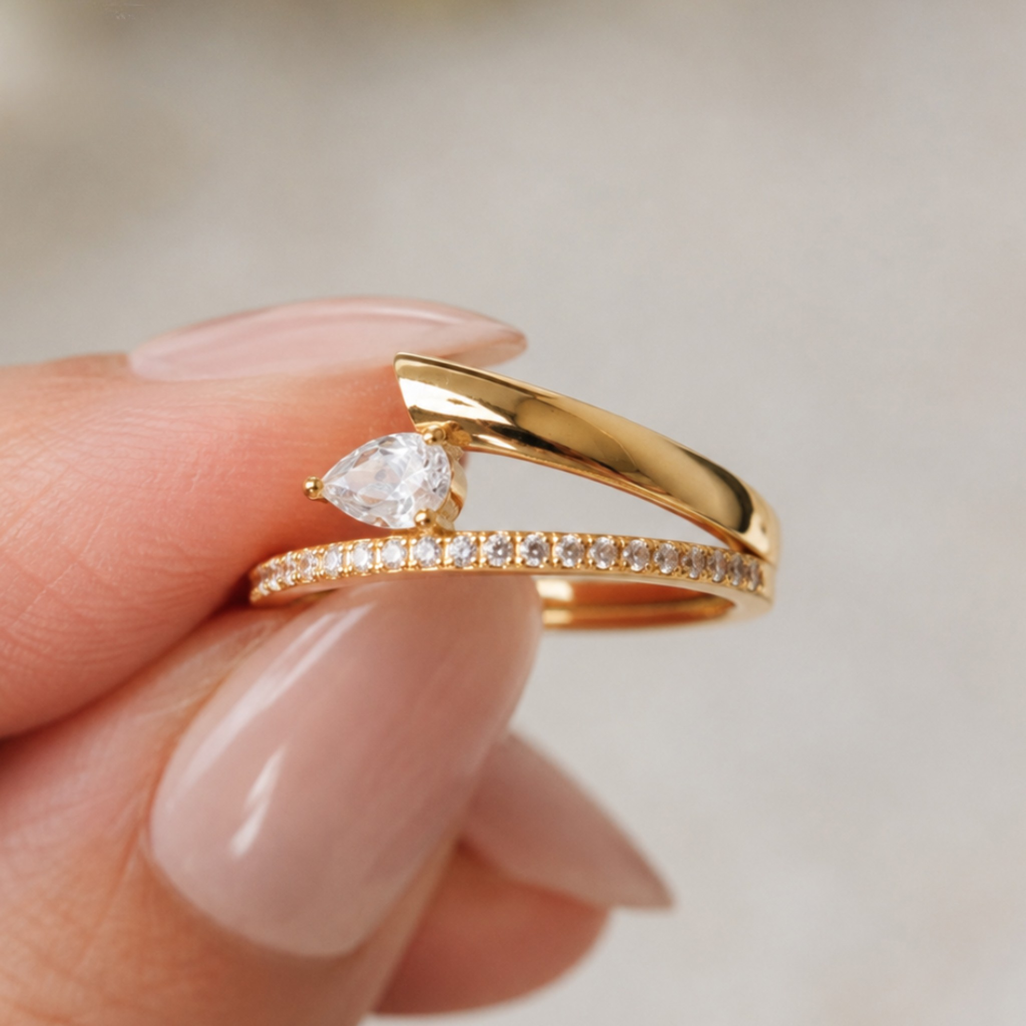 A close-up of a hand with manicured nails holding a gold ring set with a pear-cut clear gemstone and a smaller pave-studded band, against a soft light gray background.