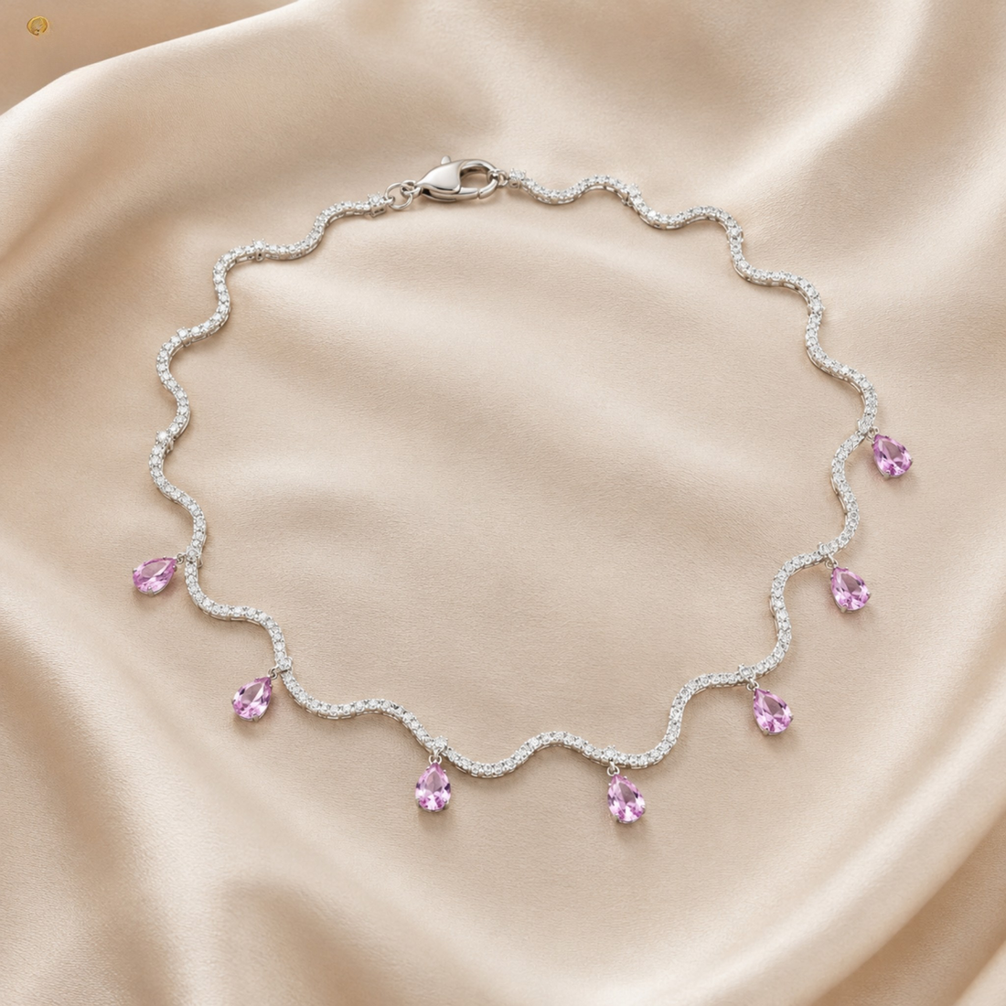 A silver-toned wavy necklace with small clear stones and dangling light pink teardrop gemstones, arranged on a soft beige satin fabric background.