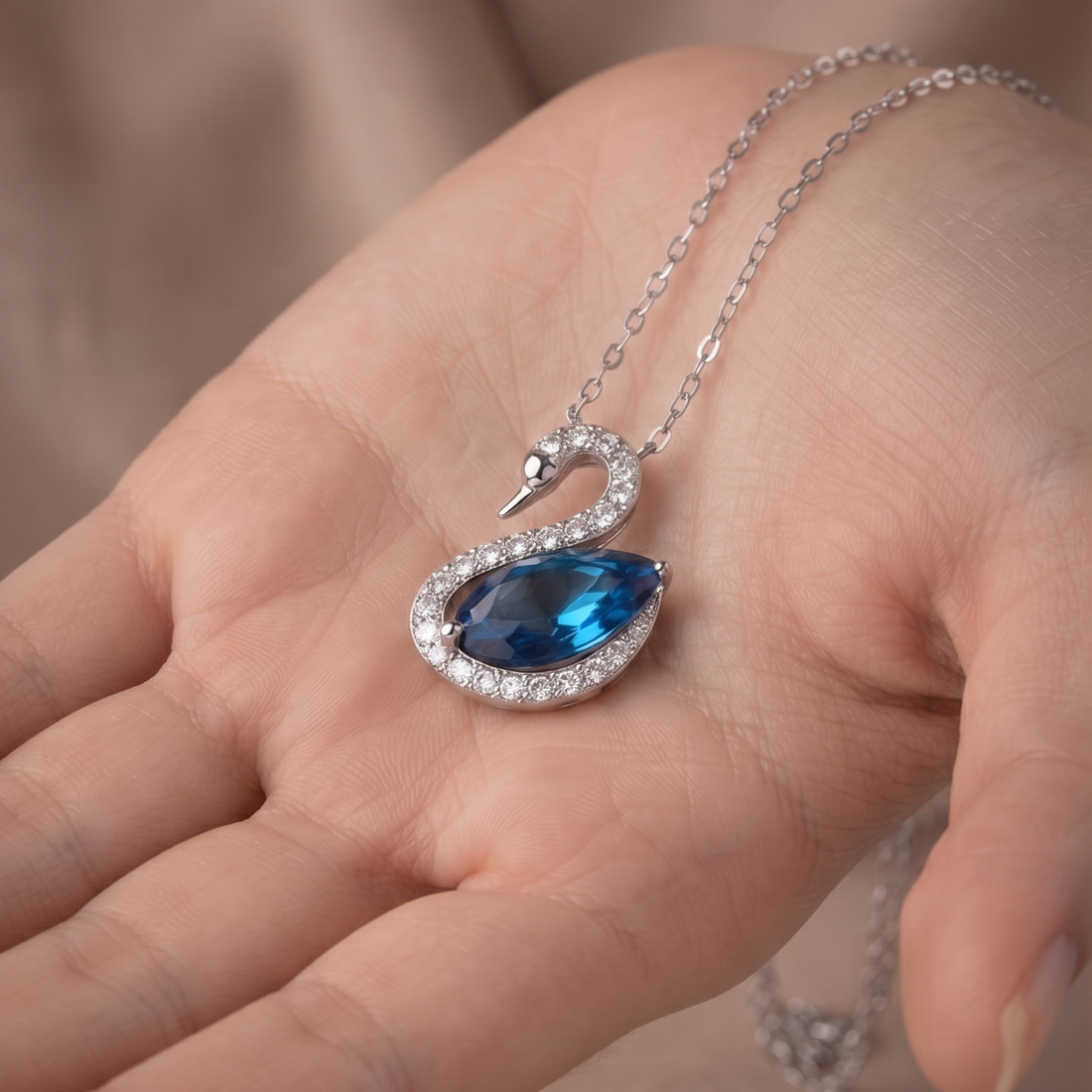 A silver-toned swan pendant necklace with a large blue teardrop gemstone and clear accent stones, displayed on an open hand.