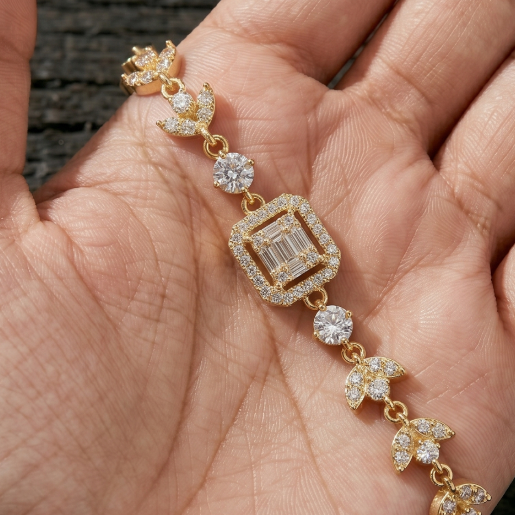 A gold-toned bracelet with clear gemstones, including a central emerald-cut stone and pavé accents, displayed on a person's palm.