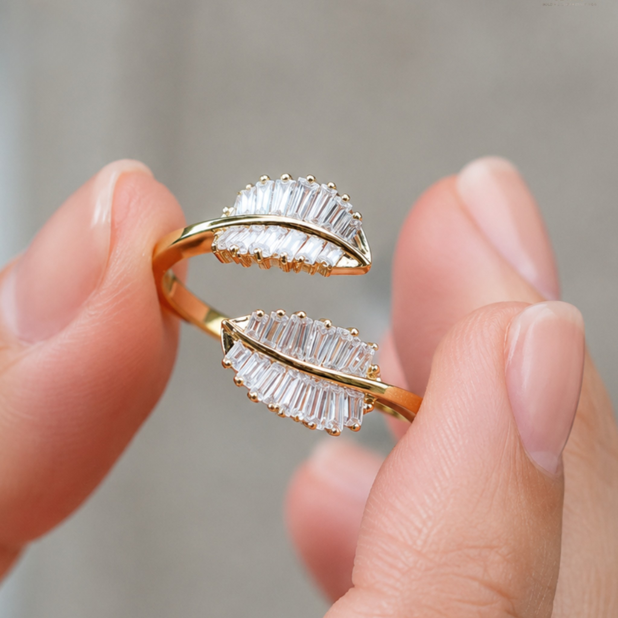 A close-up of a person's fingers holding a gold leaf-shaped ring adorned with clear baguette crystals, set against a soft, out-of-focus gray background.