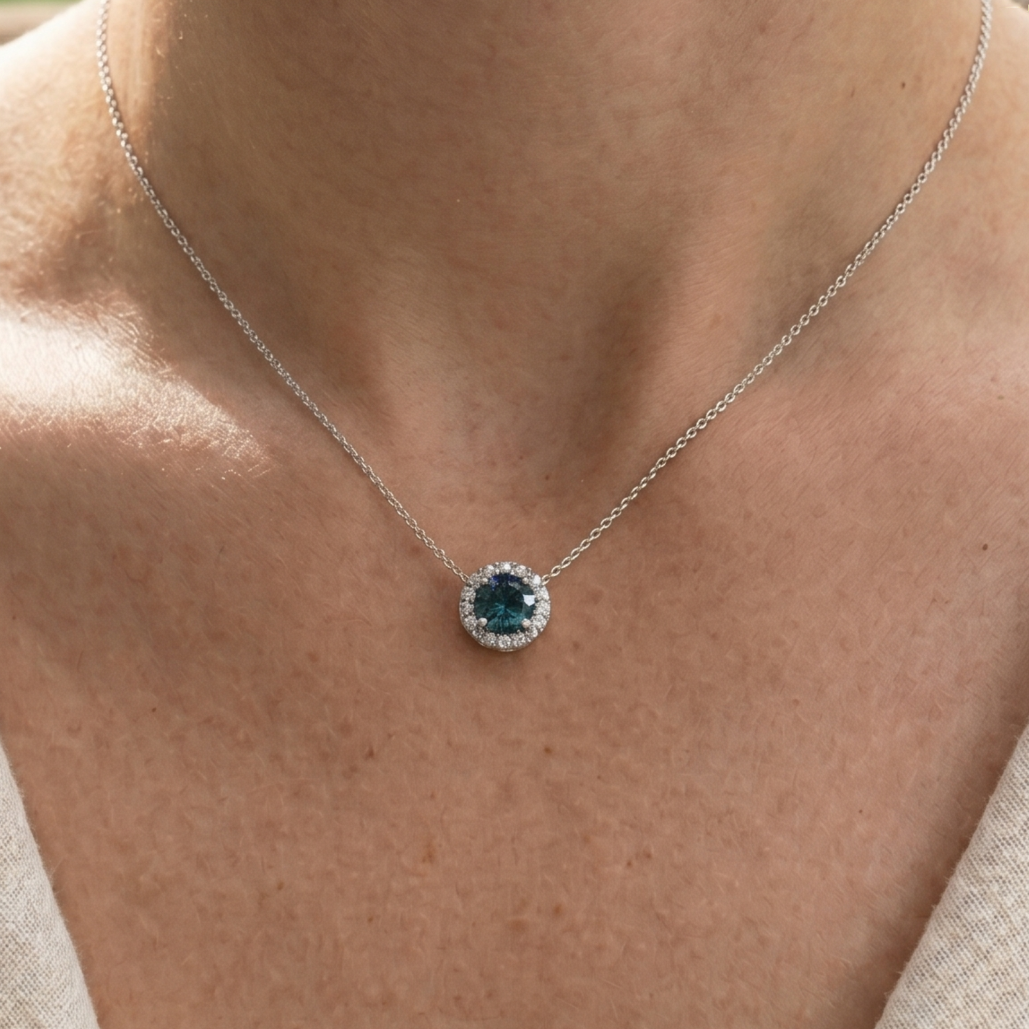 A close-up of a person's neck and upper chest wearing a delicate silver chain necklace with a round pendant. The pendant features a central deep blue gemstone surrounded by a halo of small clear stones.