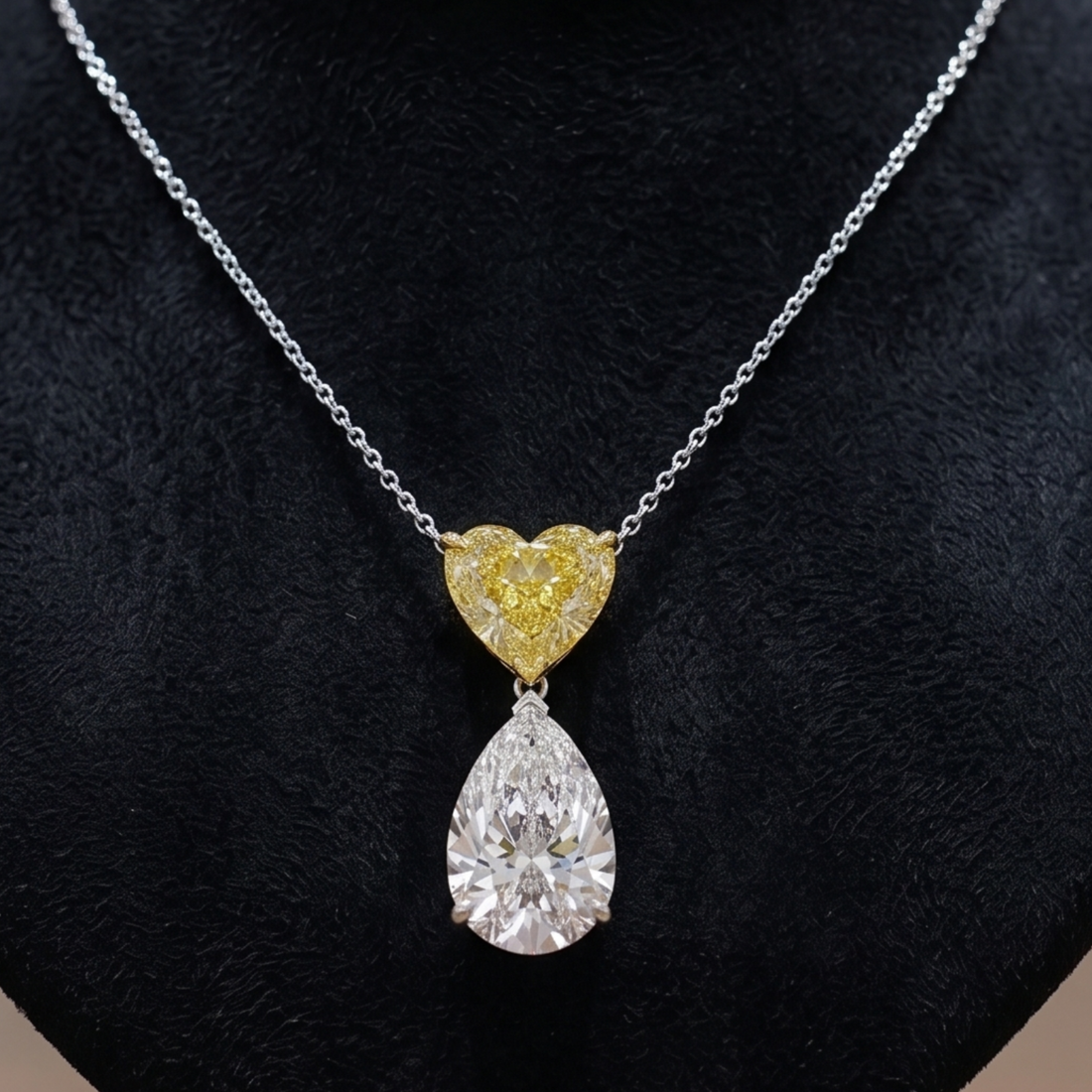 A silver chain necklace with a pendant featuring a small yellow heart-shaped diamond and a larger clear pear-shaped diamond below, displayed on a black textured bust.