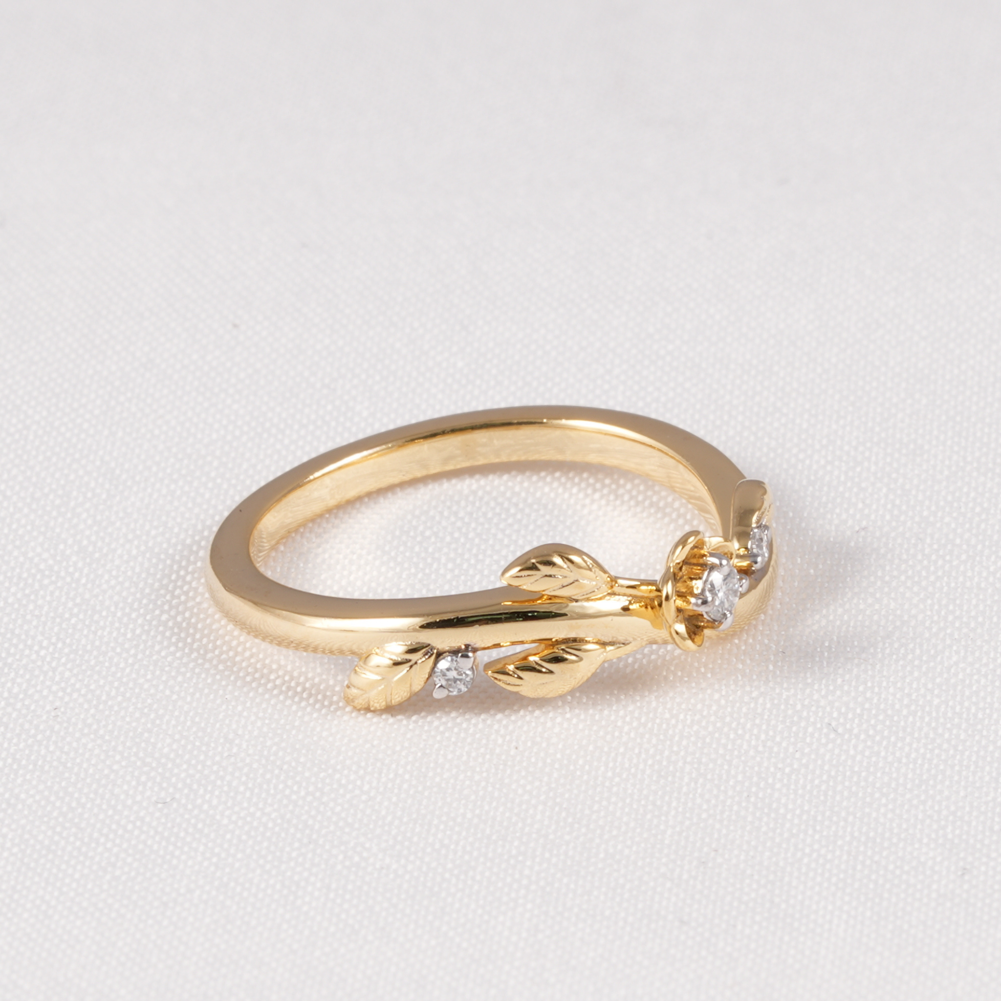 A gold ring featuring a delicate leaf and flower motif with small clear gemstones, displayed on a textured white background.