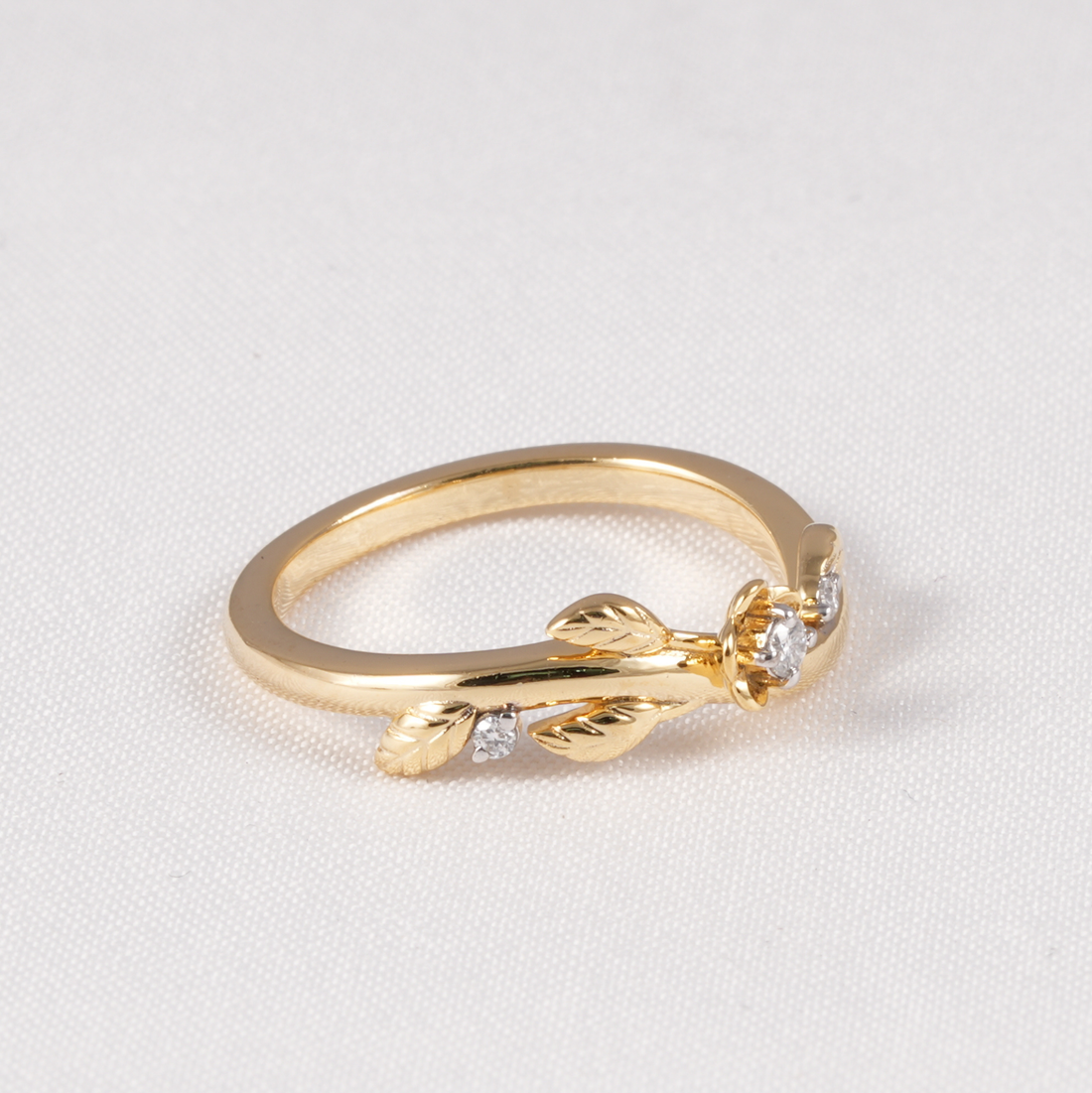 A gold ring featuring a delicate leaf and flower motif with small clear gemstones, displayed on a textured white background.
