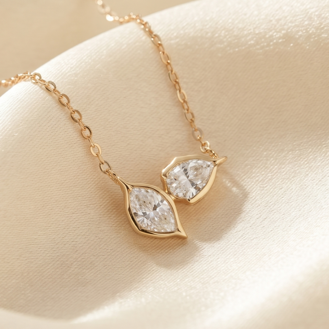A gold necklace with two marquise-cut clear gemstones in matching gold settings, displayed on a soft cream satin surface.