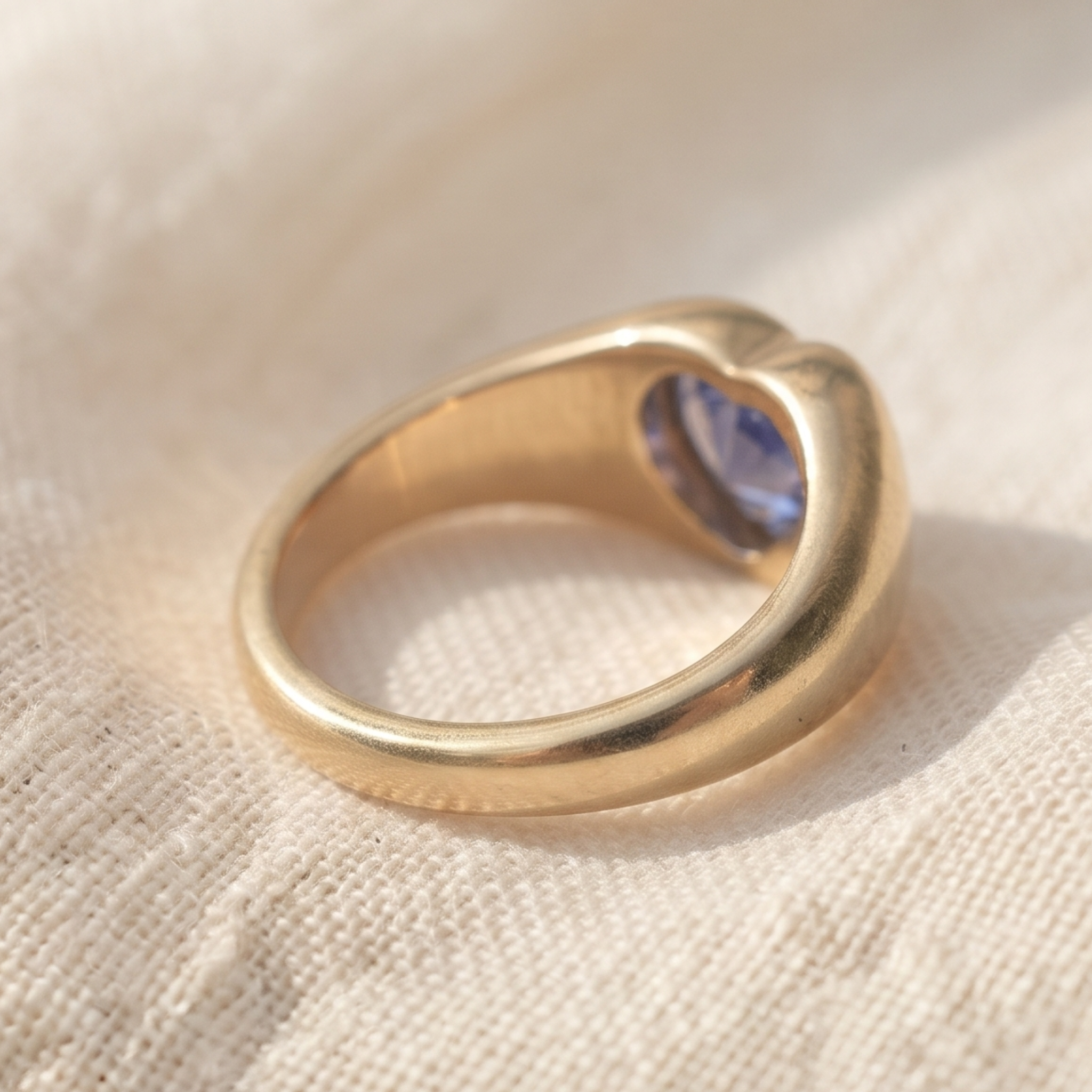 A gold ring with a heart-shaped bezel holding a round blue gemstone, resting on a textured beige fabric background.