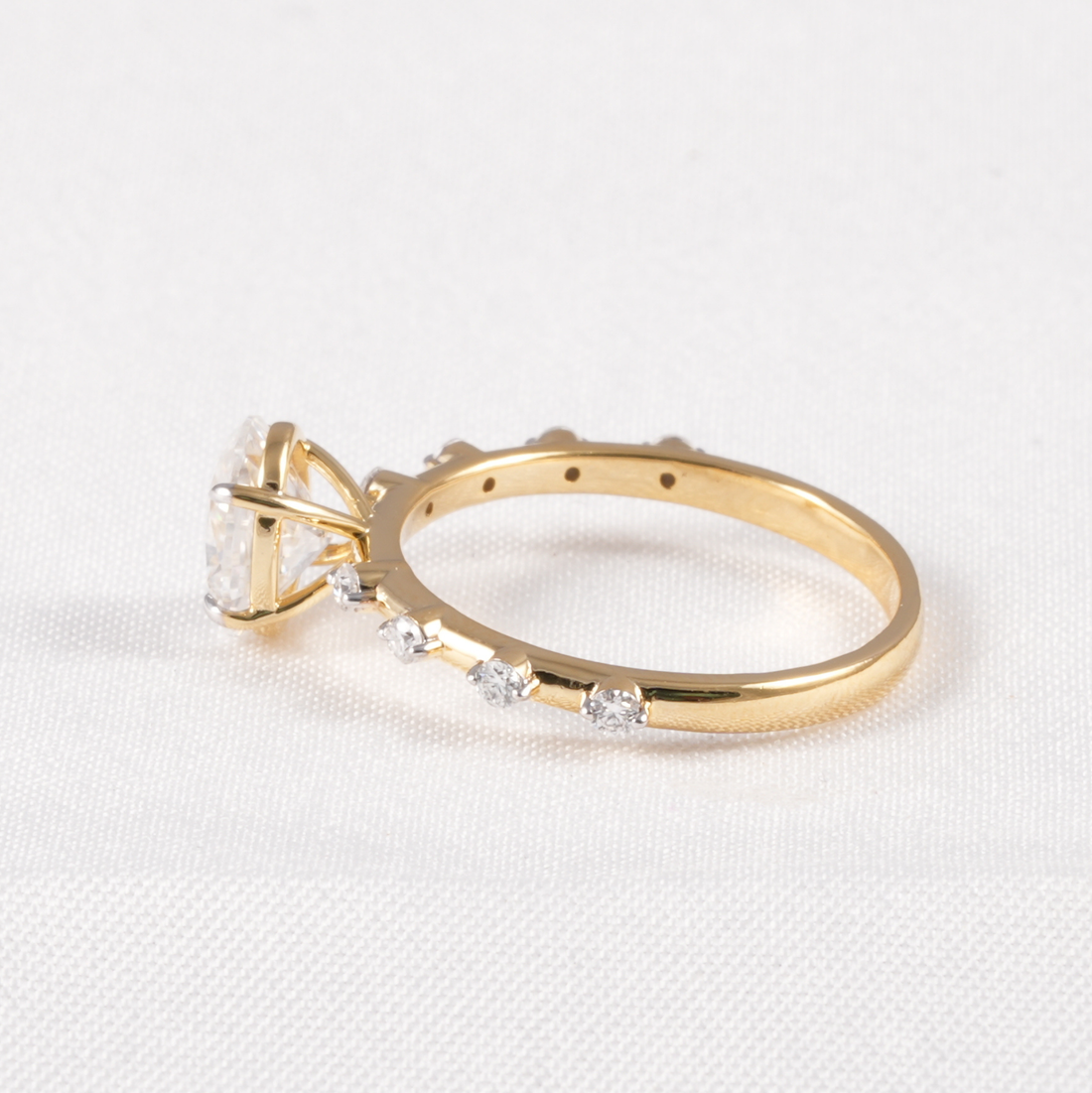 A yellow gold ring with a large clear gemstone and smaller clear stones along the band, photographed on a white textured fabric background.