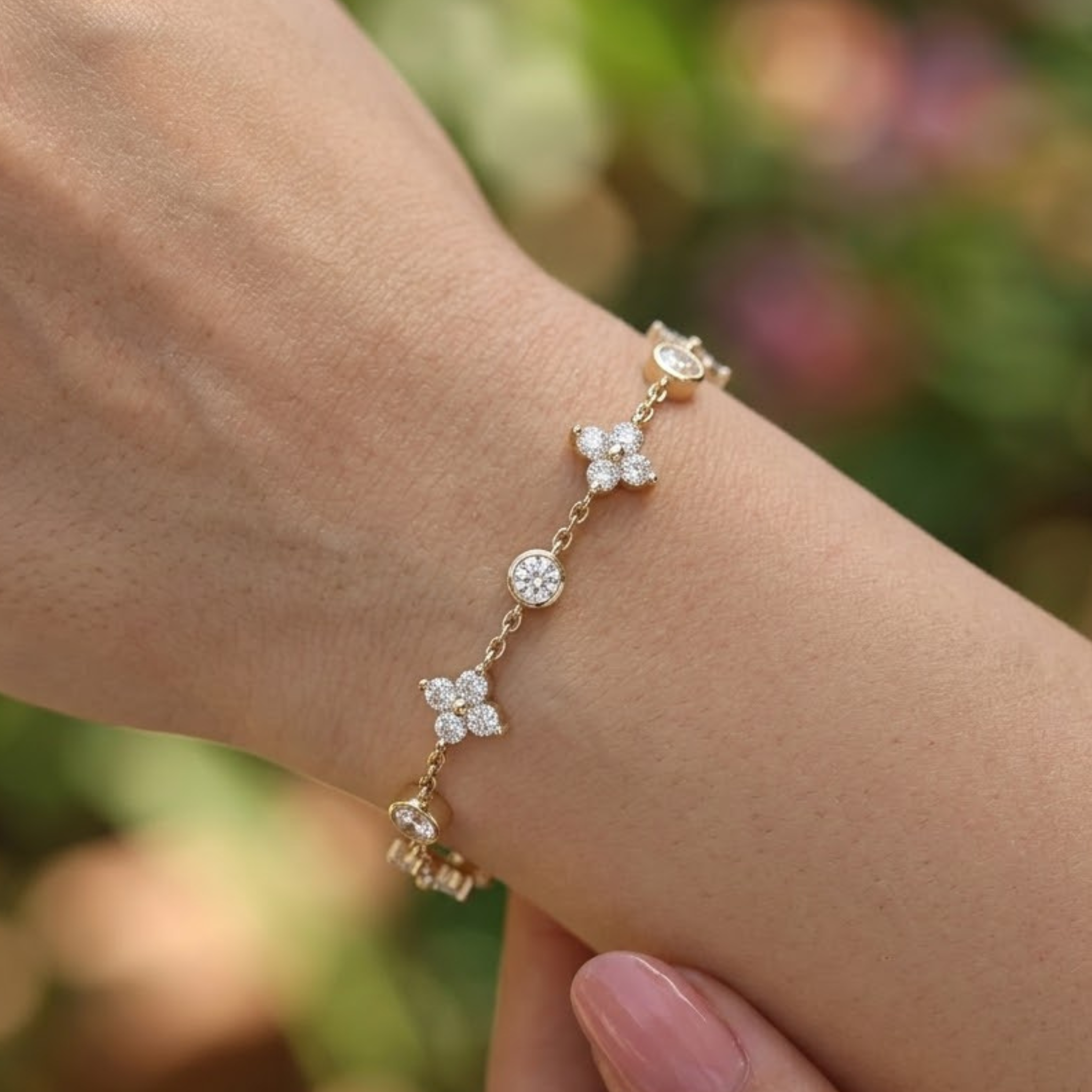 A delicate gold chain bracelet with four floral clusters set with clear stones, shown worn on a person's wrist against a blurred green and pink outdoor background.