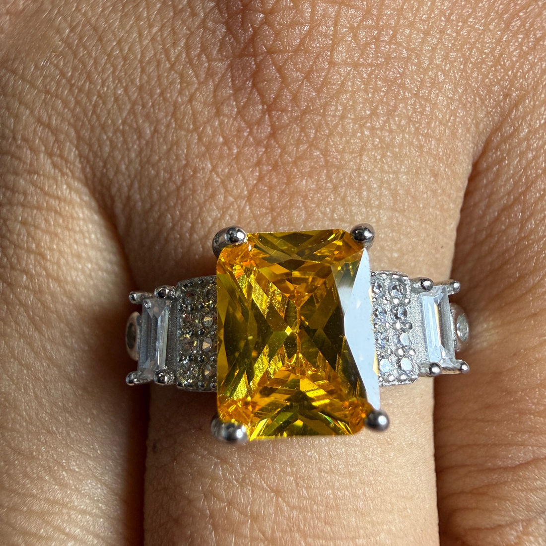 A silver ring featuring a large rectangular yellow gemstone with baguette and round side stones, shown worn on a finger.