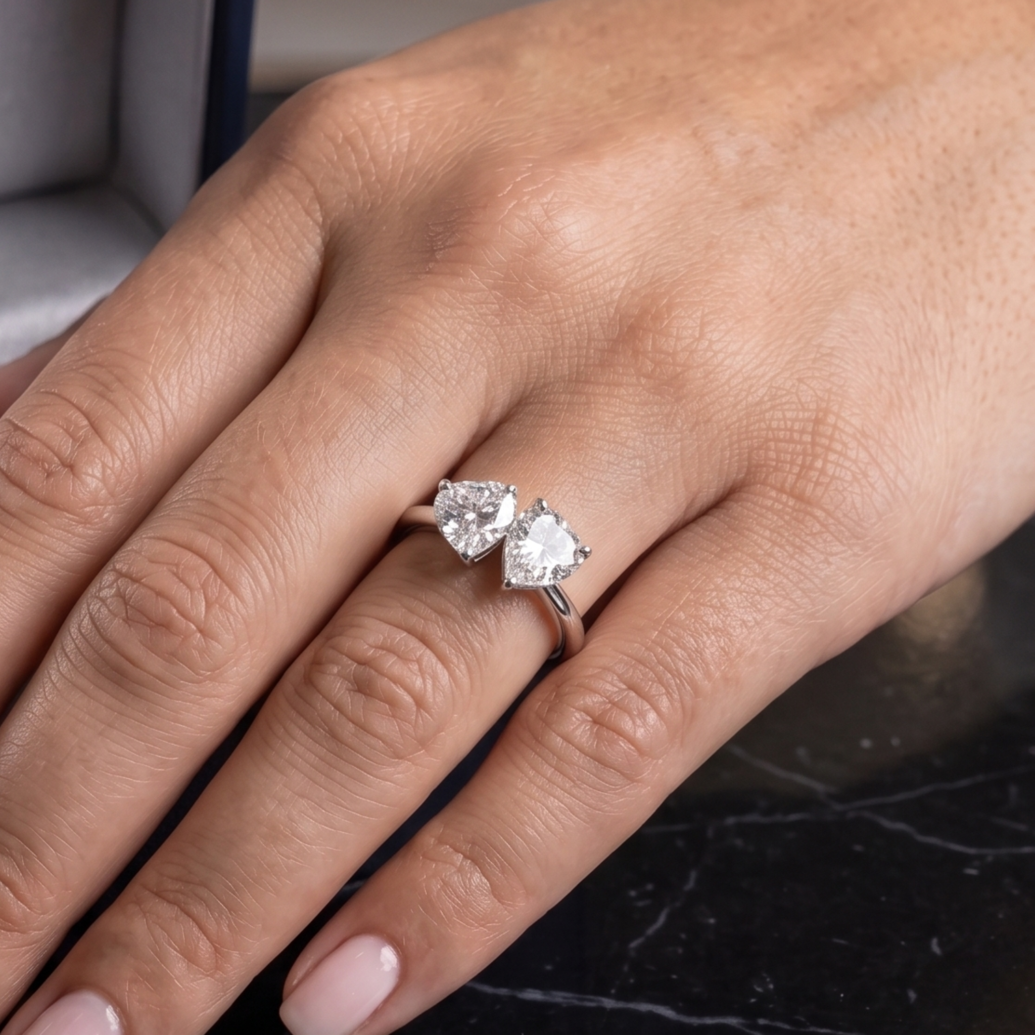 A close-up of a hand wearing a silver ring with two heart-shaped clear gemstones, set against a dark marble surface.