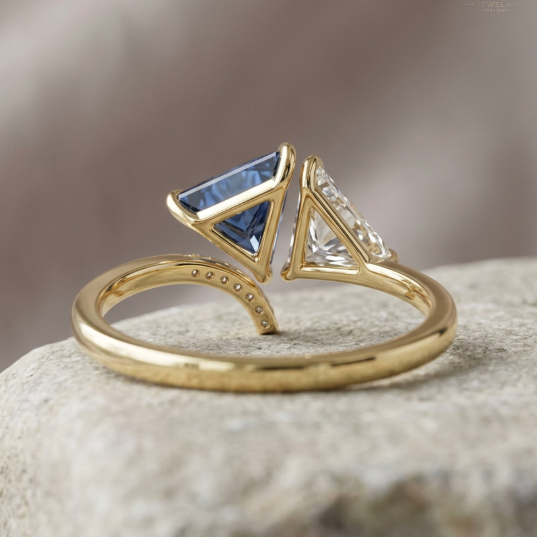 A gold ring with a split triangular design, featuring a blue gemstone on one side and a clear gemstone on the other, resting on a textured stone surface.