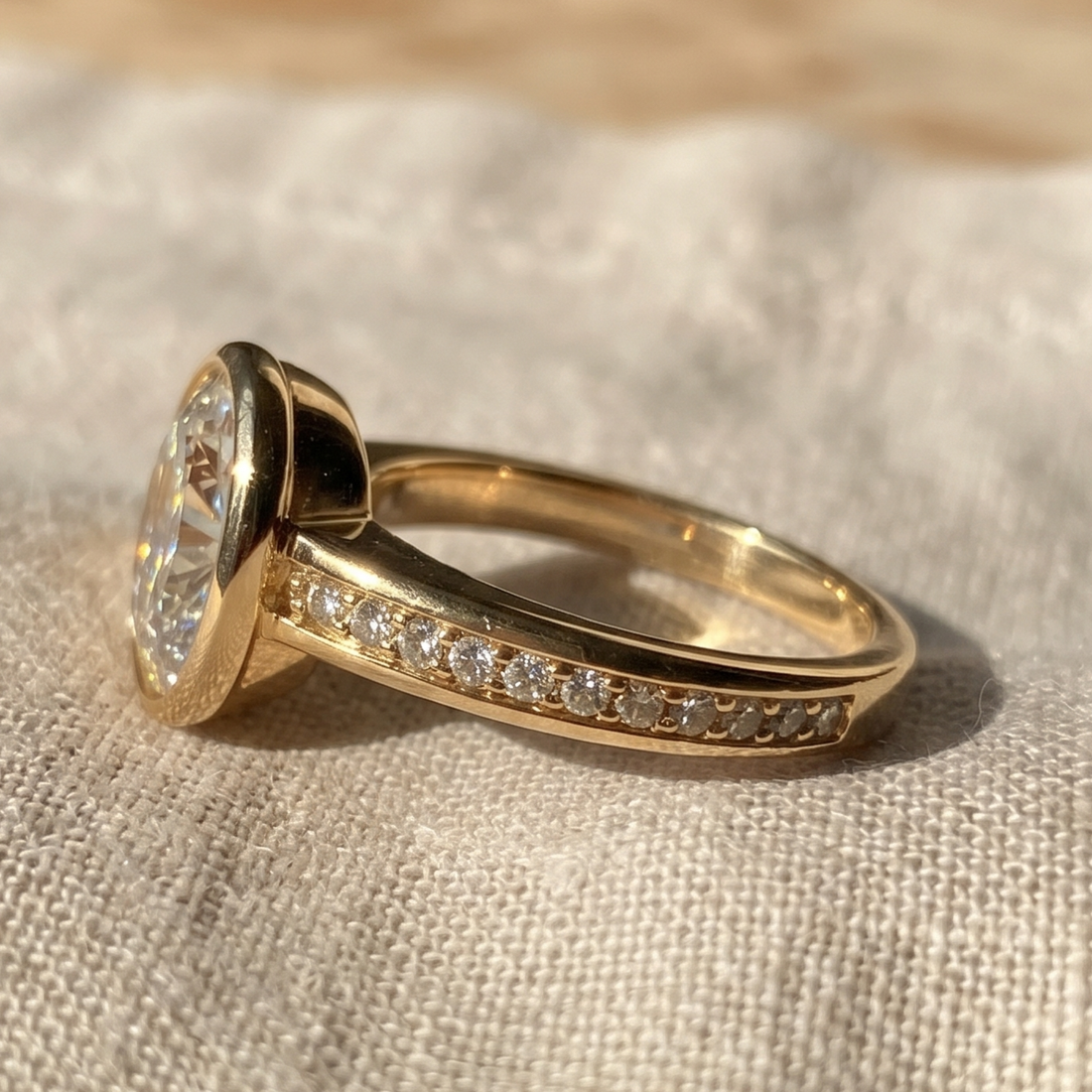 A gold ring with a large oval clear gemstone and a pavé band of small clear stones, resting on a textured beige fabric surface.