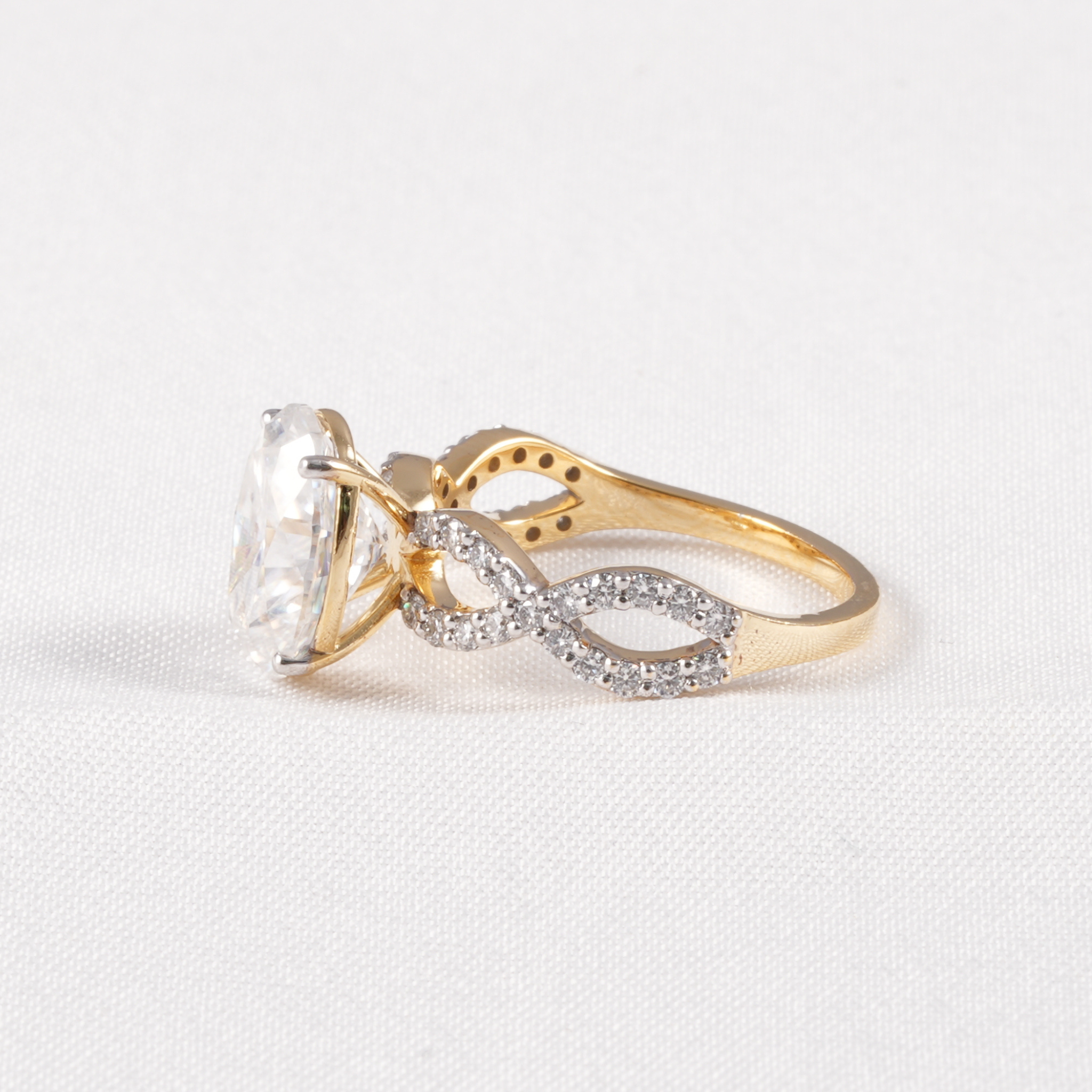 A gold ring featuring a large clear oval-cut center stone and an infinity-shaped band paved with small clear stones, shown on a light textured background.