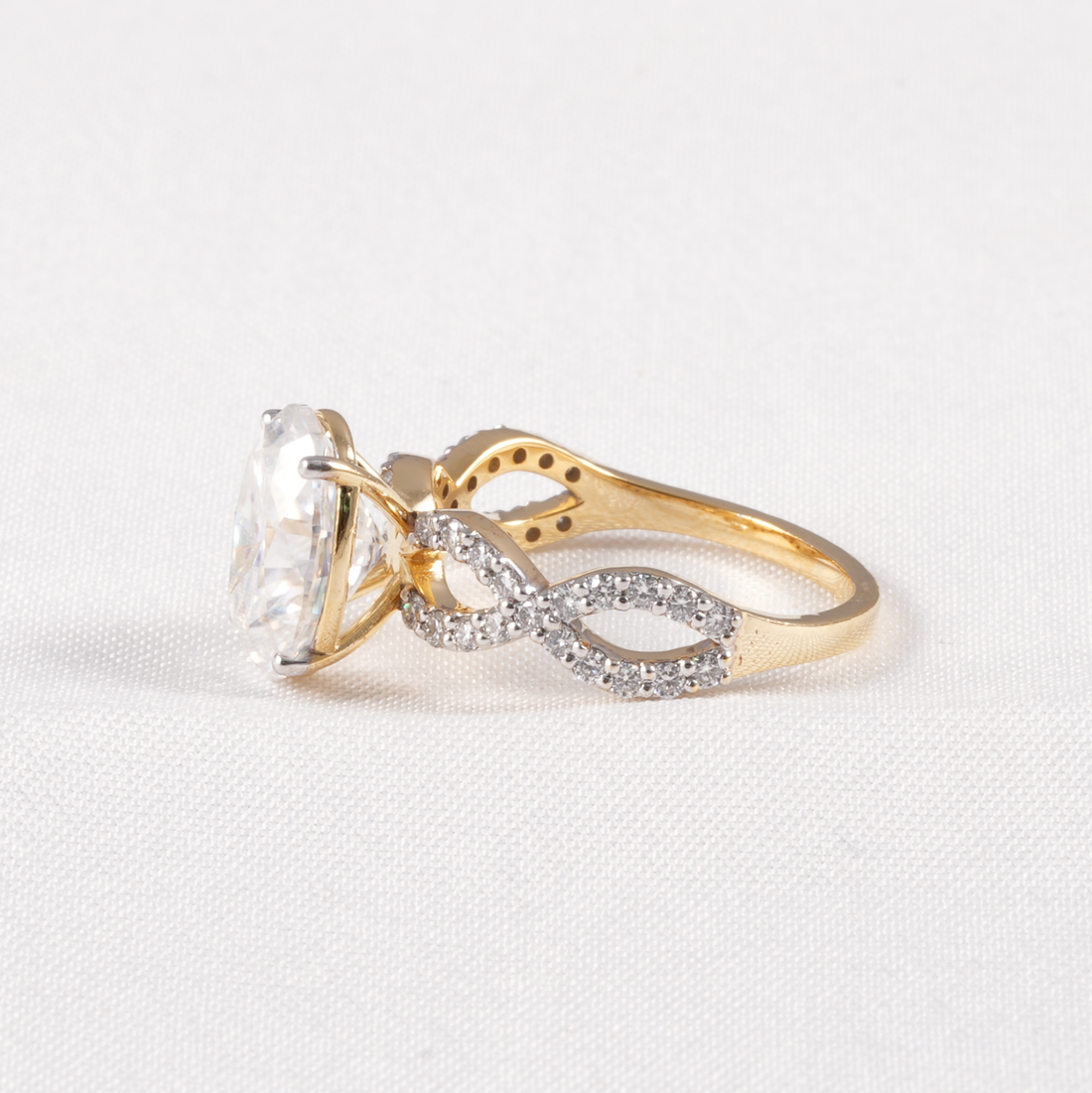 A gold ring featuring a large clear oval-cut center stone and an infinity-shaped band paved with small clear stones, shown on a light textured background.