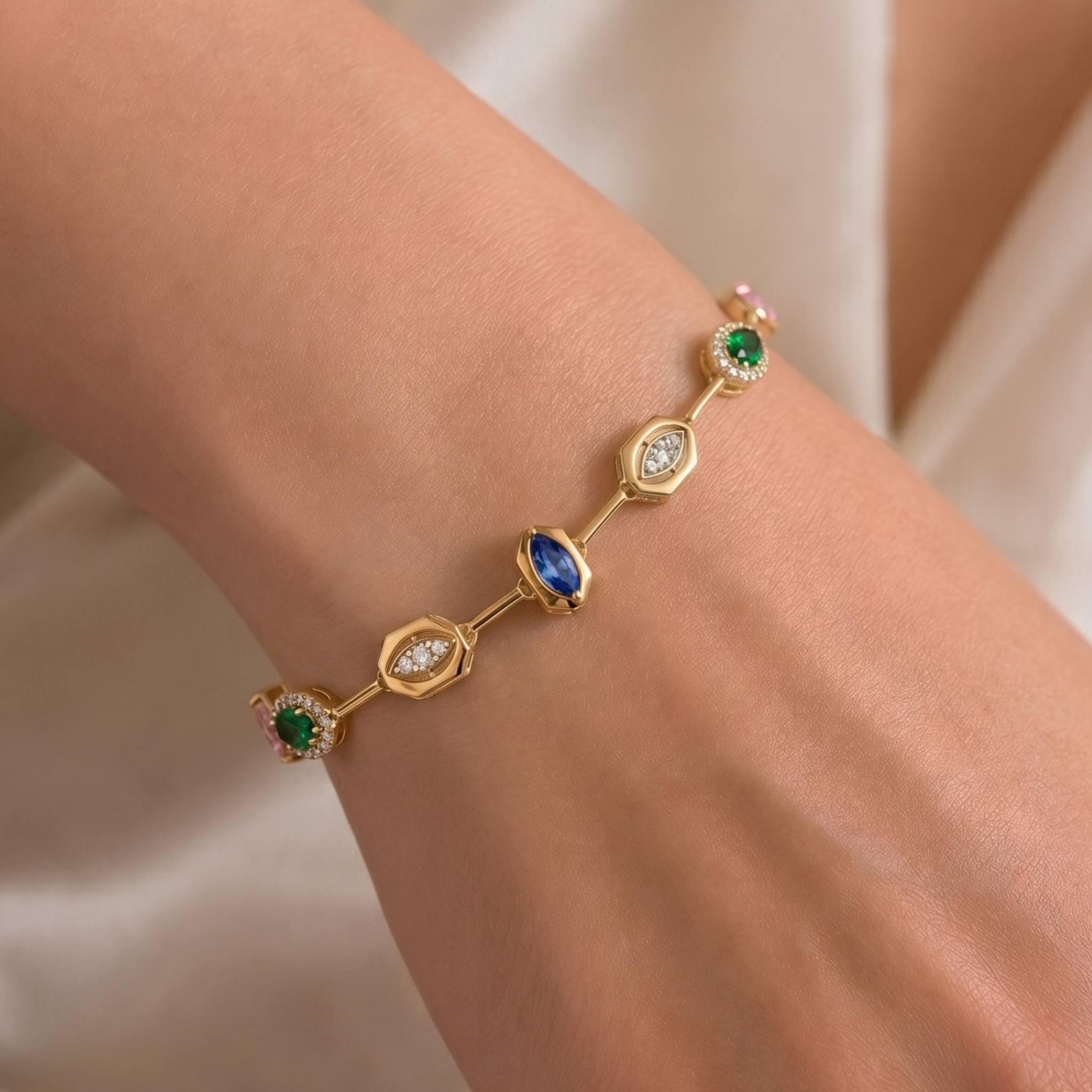A gold bracelet with green and blue gemstones and clear accent stones, shown worn on a person's wrist against a soft cream fabric background.