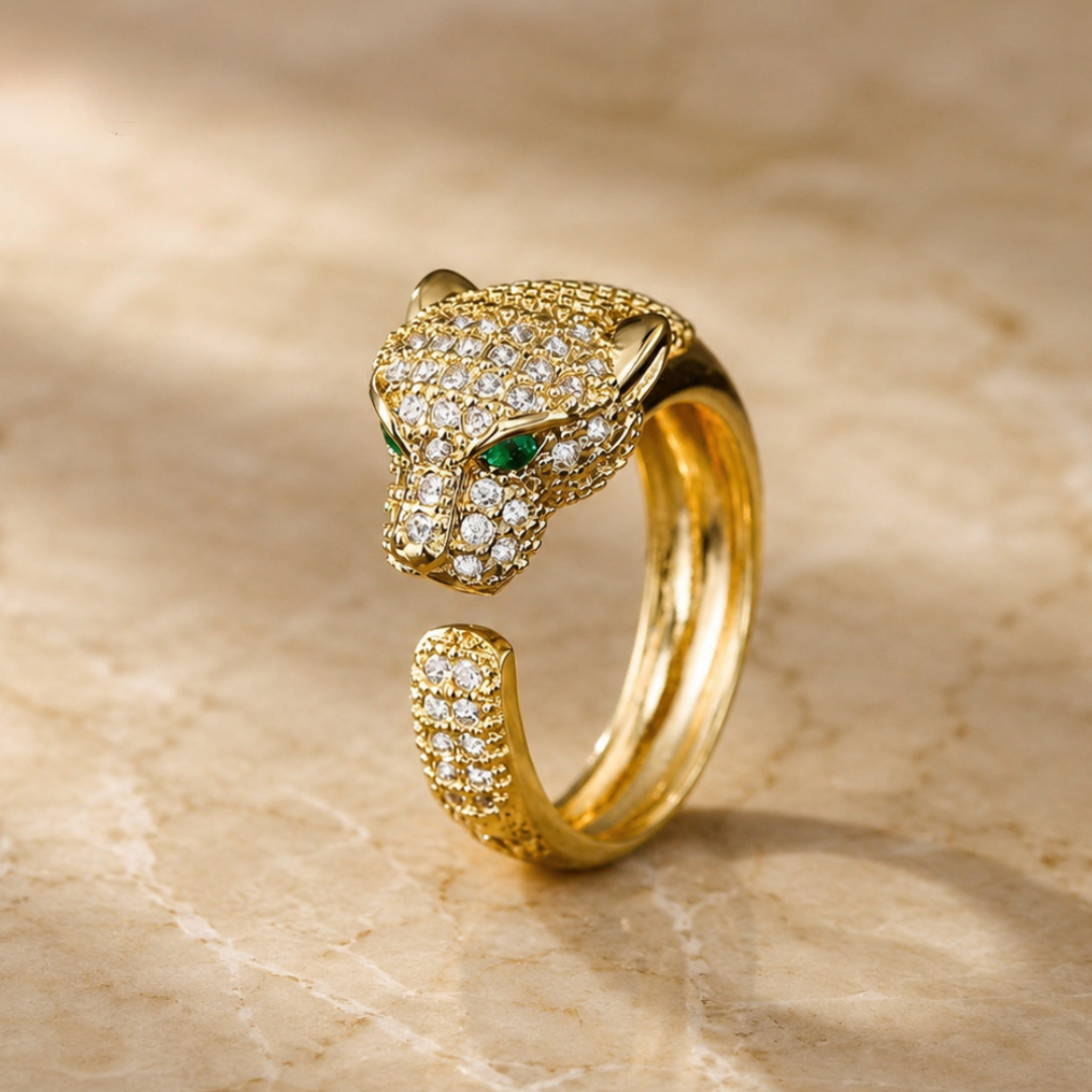 A gold open-bend ring shaped like a panther head, adorned with pavé-set clear stones and small green stone eyes, photographed on a beige stone surface.