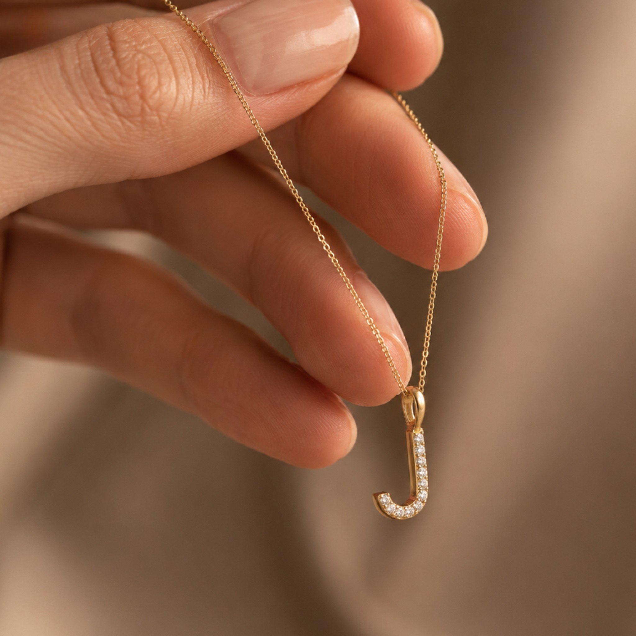 A close-up of a hand holding a delicate gold chain necklace with a small pendant shaped like the letter 'J', accented with small clear stones, set against a soft, warm beige background.
