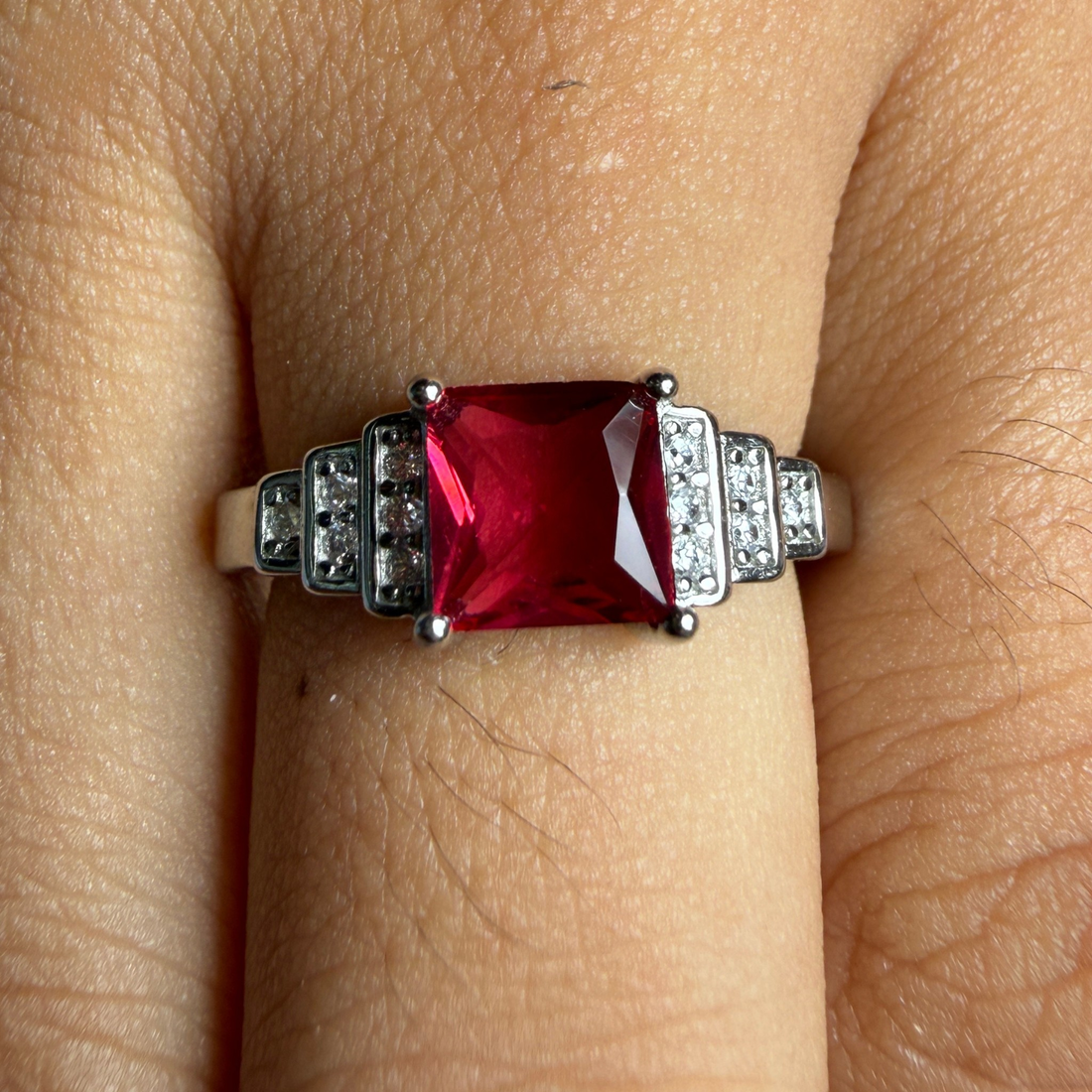 A close-up of a ring worn on a finger, featuring a rectangular red gemstone in a silver-toned setting with small clear accent stones on either side.