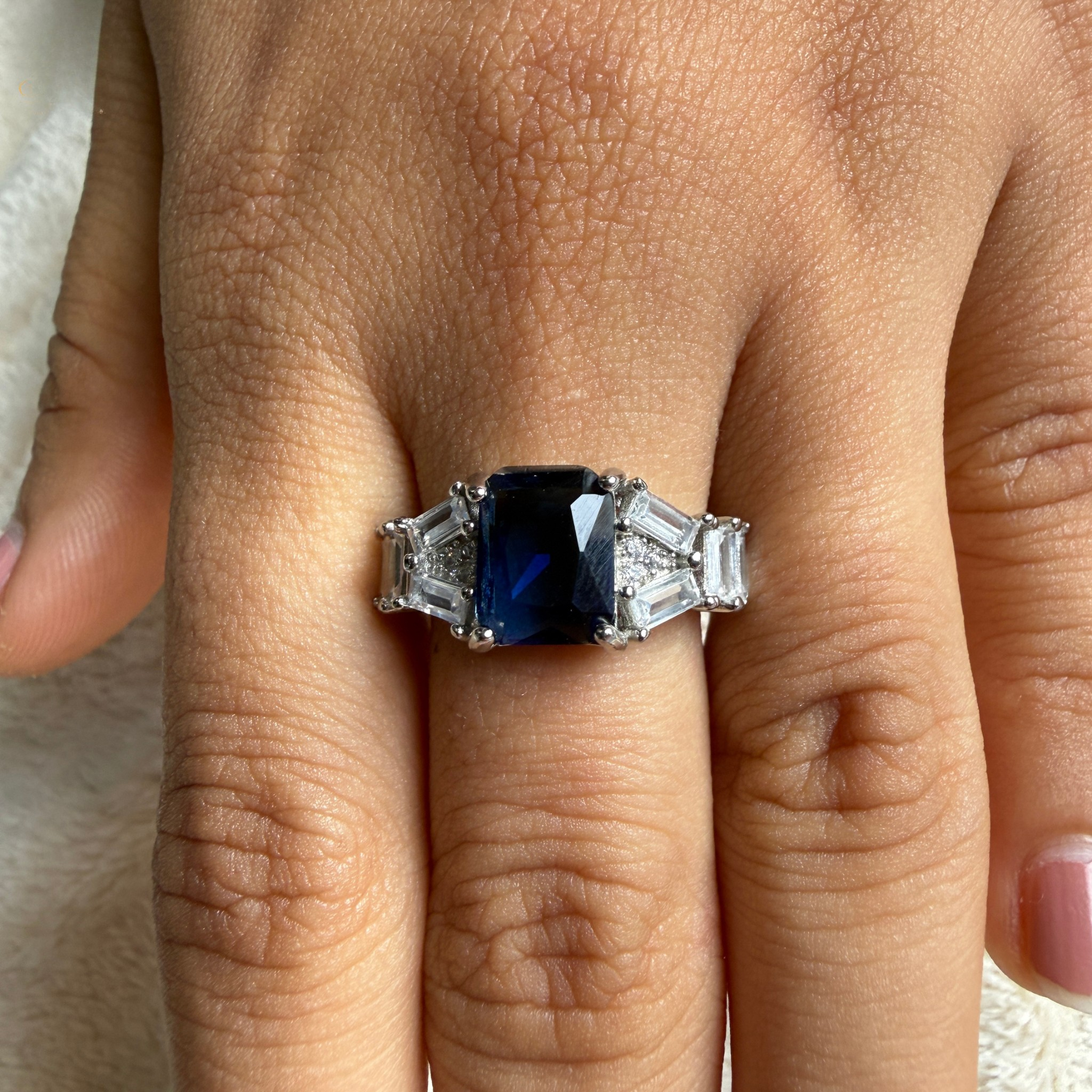 A silver ring featuring a rectangular dark blue sapphire center stone and baguette-cut side stones, shown worn on a person's finger.