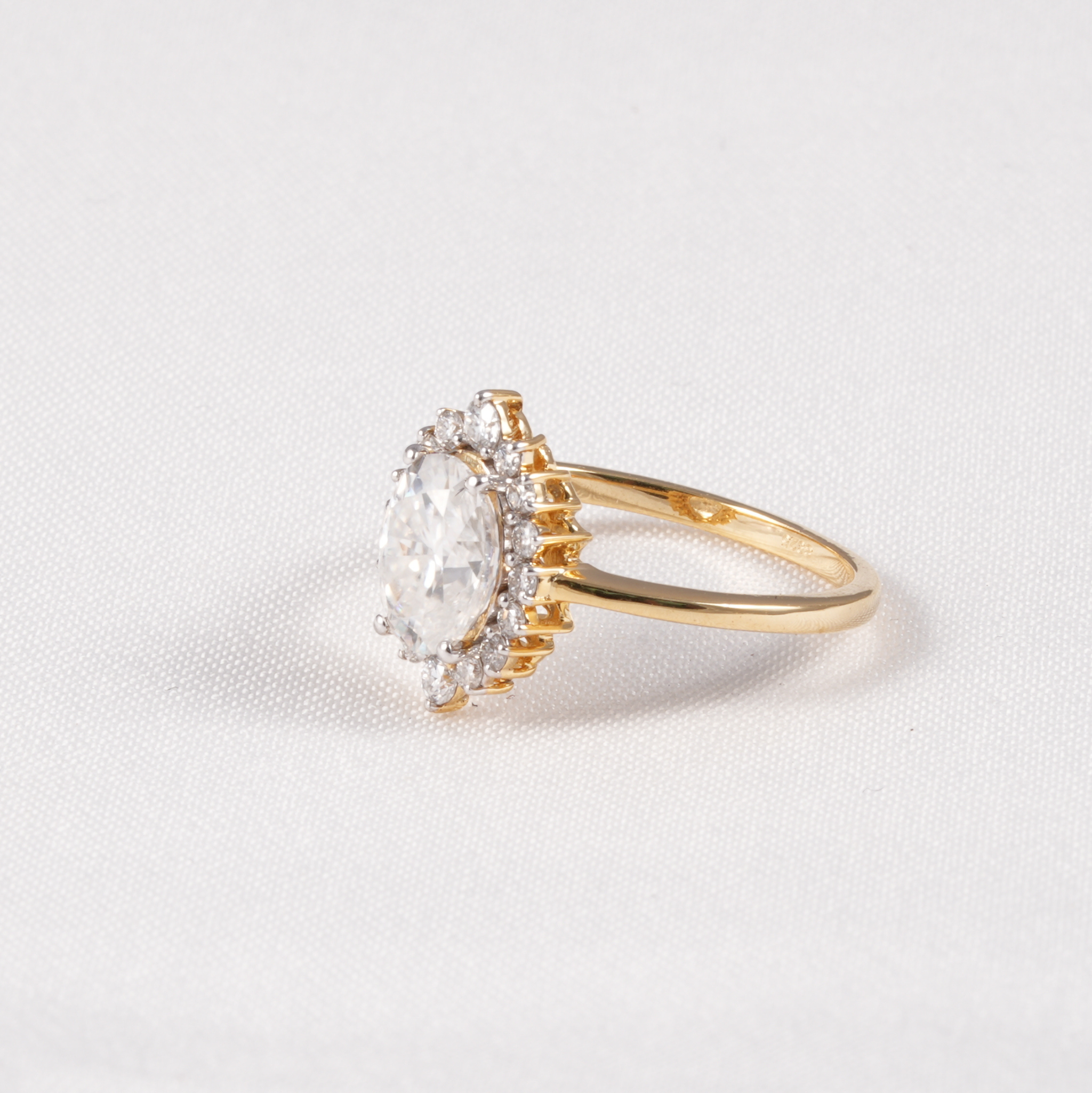 A yellow gold ring featuring a large oval-cut clear gemstone surrounded by a halo of smaller clear stones, set on a textured white background.