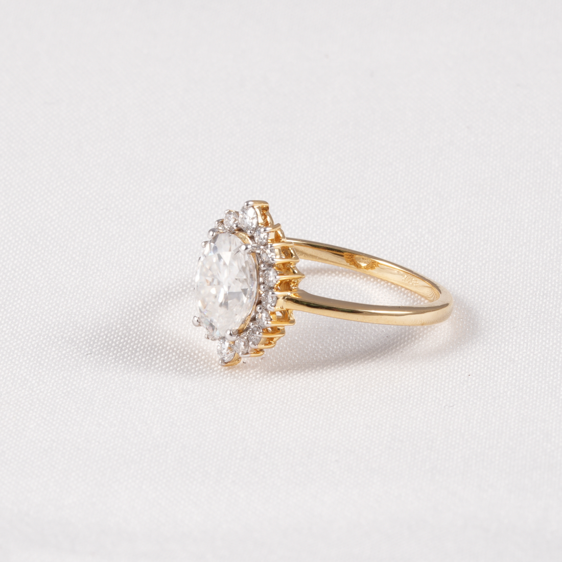 A yellow gold ring featuring a large oval-cut clear gemstone surrounded by a halo of smaller clear stones, set on a textured white background.