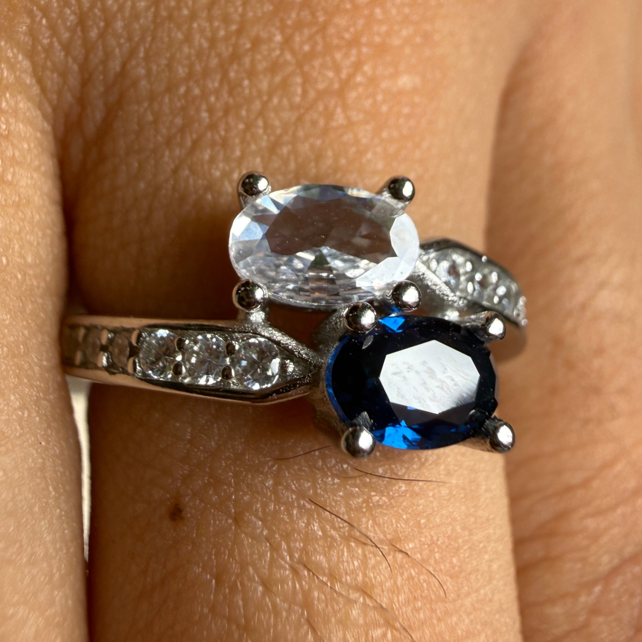 A close-up of a silver ring worn on a finger, featuring an oval clear gemstone on top, a faceted dark blue gemstone below it, and small clear stones along the band.