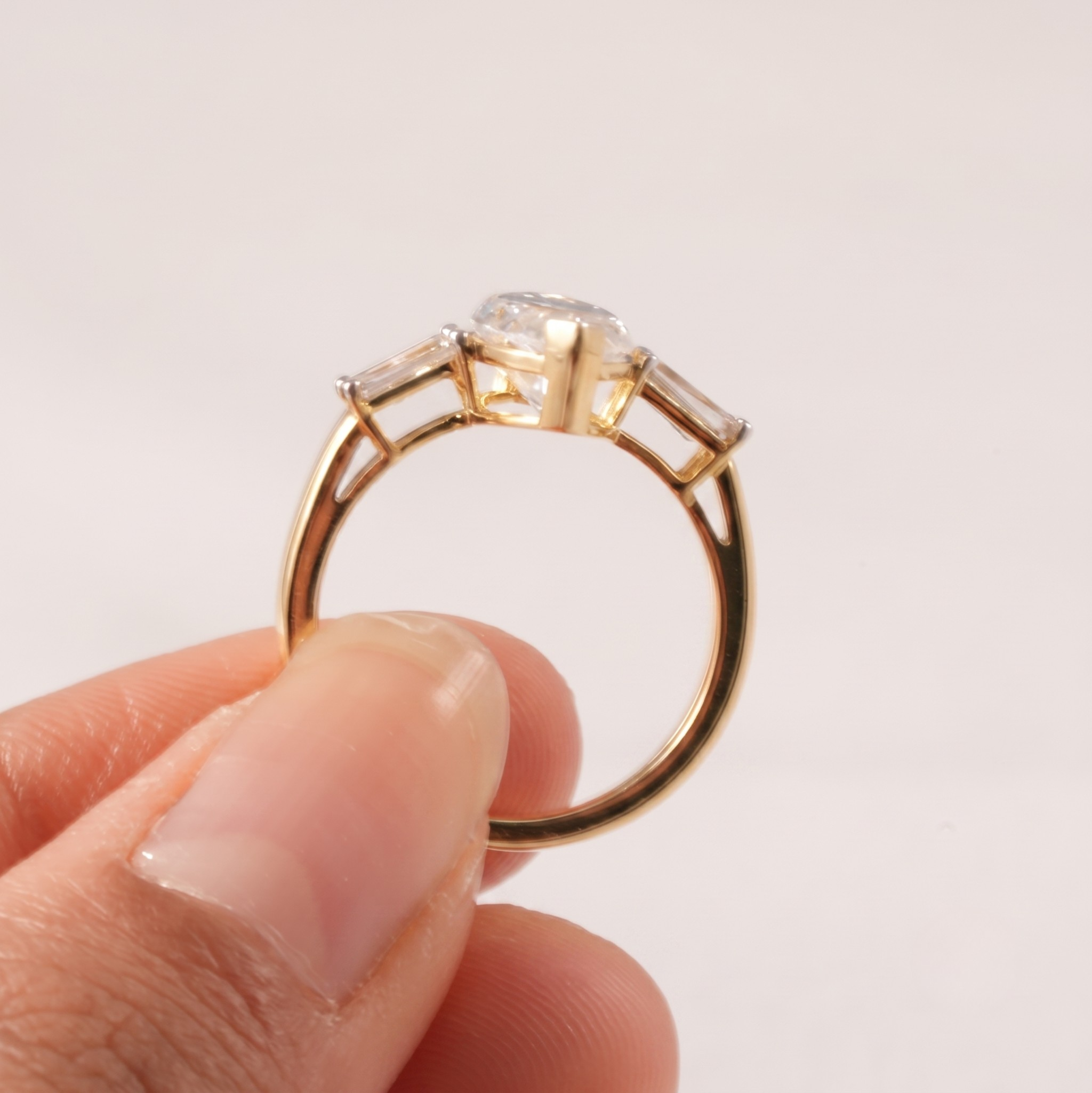 A gold ring with a large round clear center stone and two small rectangular side stones, held between fingers against a plain light background.