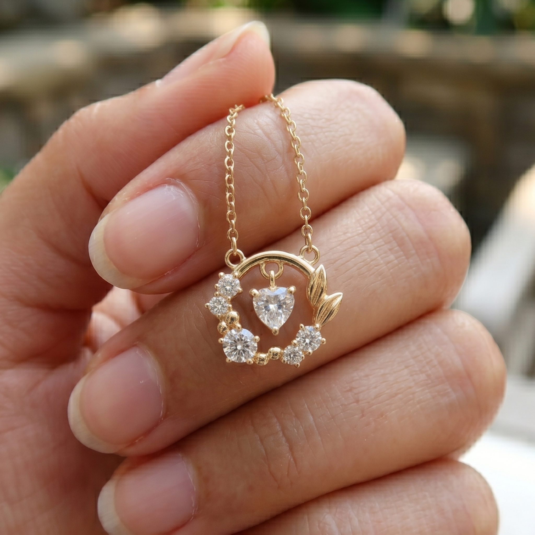 A gold necklace with a heart-shaped clear gemstone and small clear stones, held in a person's hand against a blurred outdoor background.