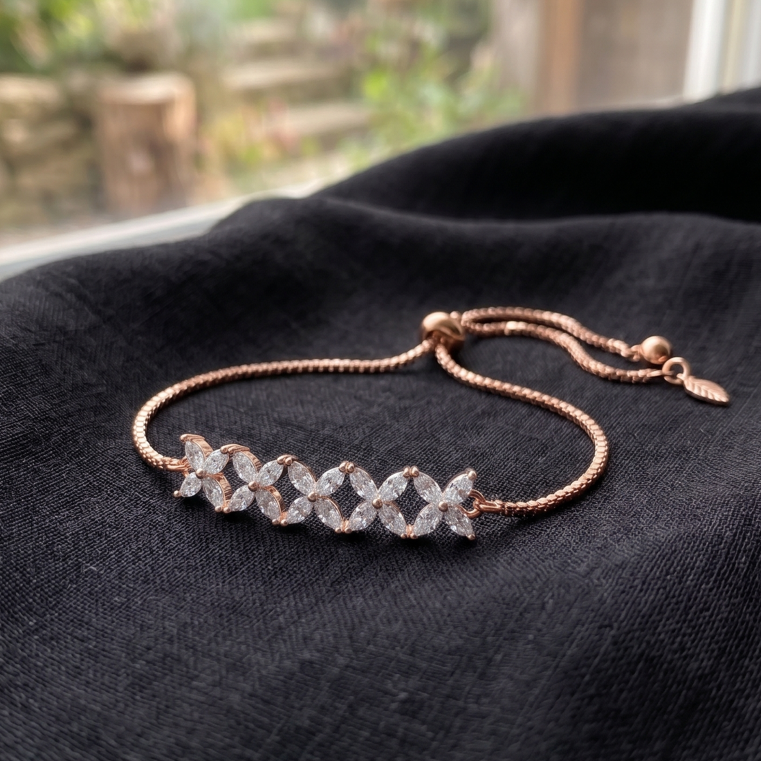 A rose gold chain bracelet with a central cluster of clear, sparkling marquise-cut gemstones, displayed on a textured black fabric surface.