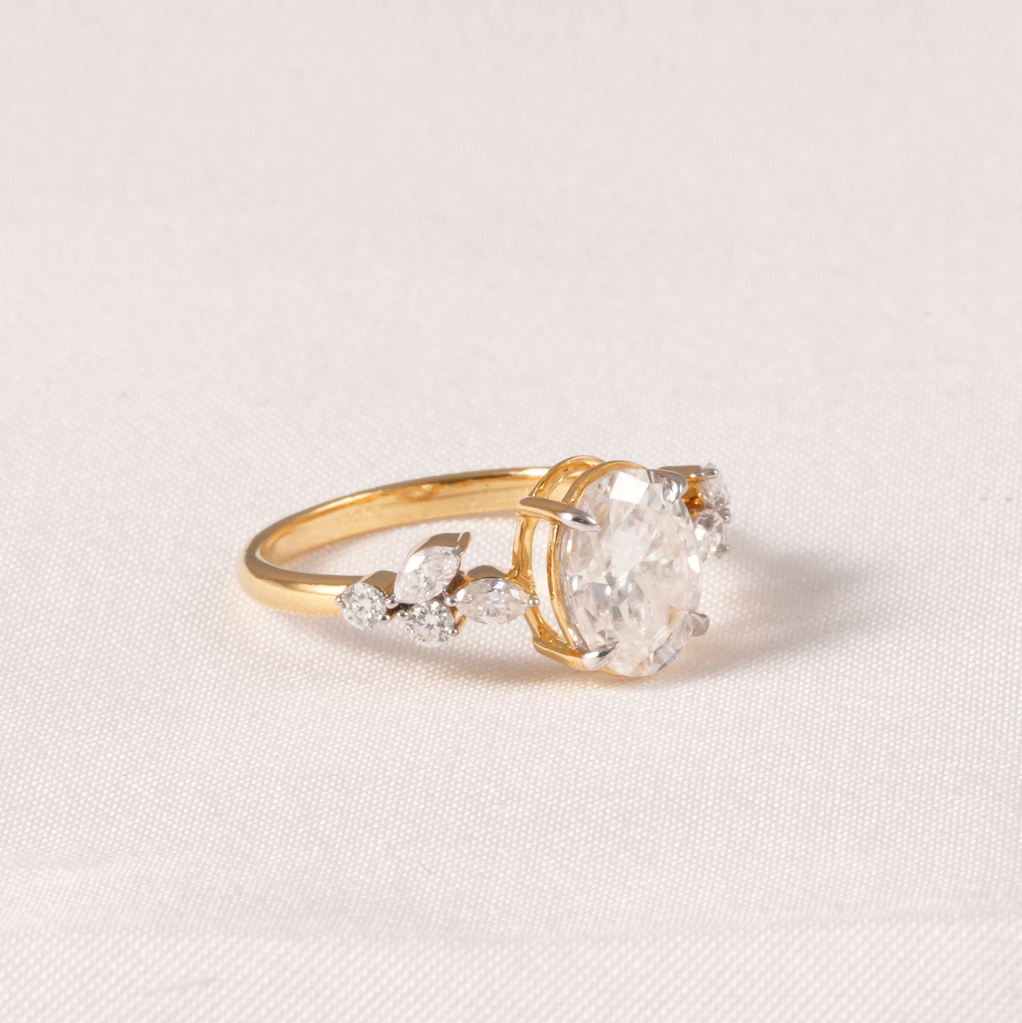 A yellow gold ring featuring a large oval clear gemstone in a prong setting, accented by smaller marquise and round clear stones along the band, set against a light beige textured fabric background.