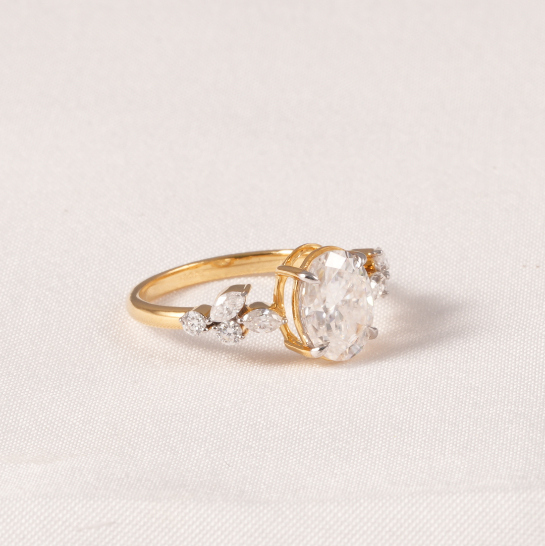 A yellow gold ring featuring a large oval clear gemstone in a prong setting, accented by smaller marquise and round clear stones along the band, set against a light beige textured fabric background.