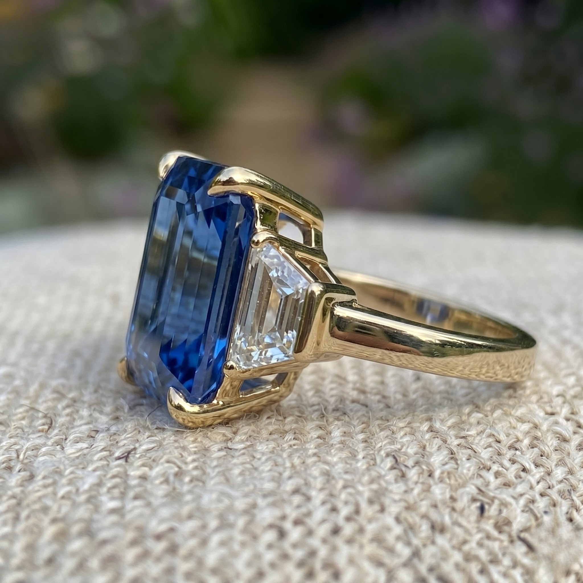 A yellow gold ring featuring a large rectangular blue sapphire gemstone and a smaller clear baguette-cut side stone, resting on a textured beige woven surface with a blurred green garden background.