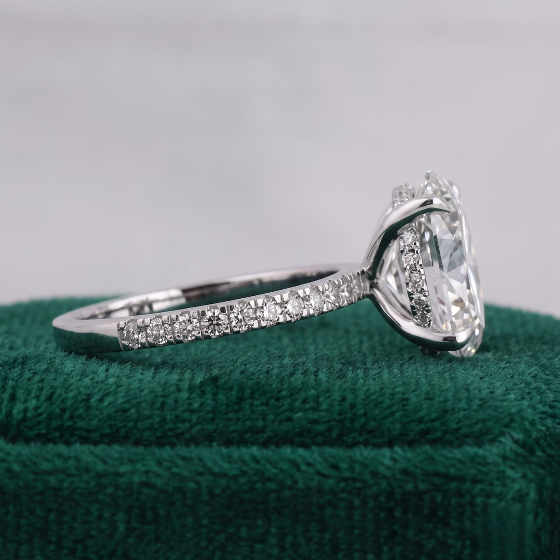 A silver-toned diamond ring with a large clear heart-shaped center stone and pavé-set diamonds along the band, displayed on a textured dark green velvet surface against a soft light gray background.