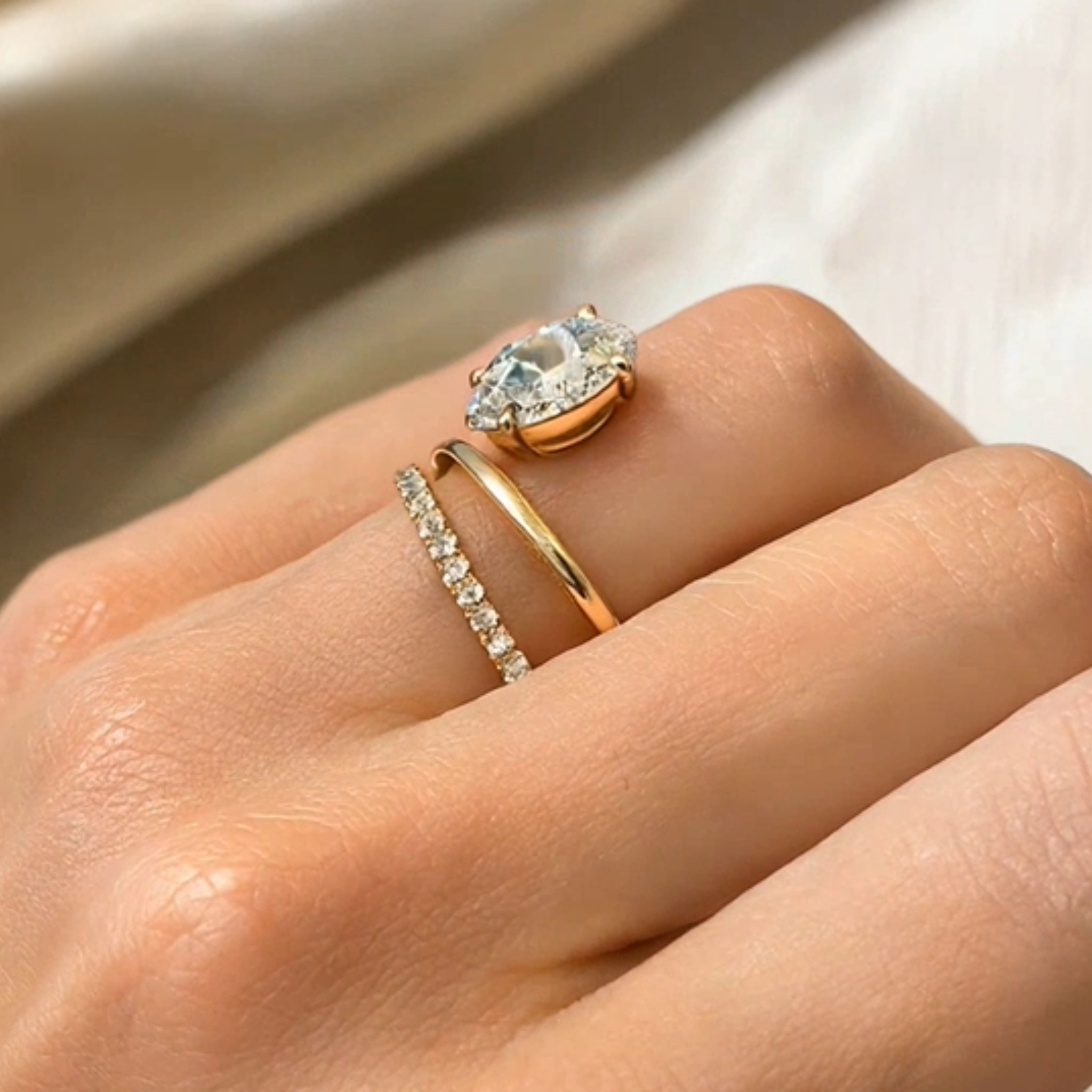 A close-up of a hand wearing two gold rings: one with a marquise-cut clear gemstone and the other a delicate pavé band, against a soft beige fabric background.