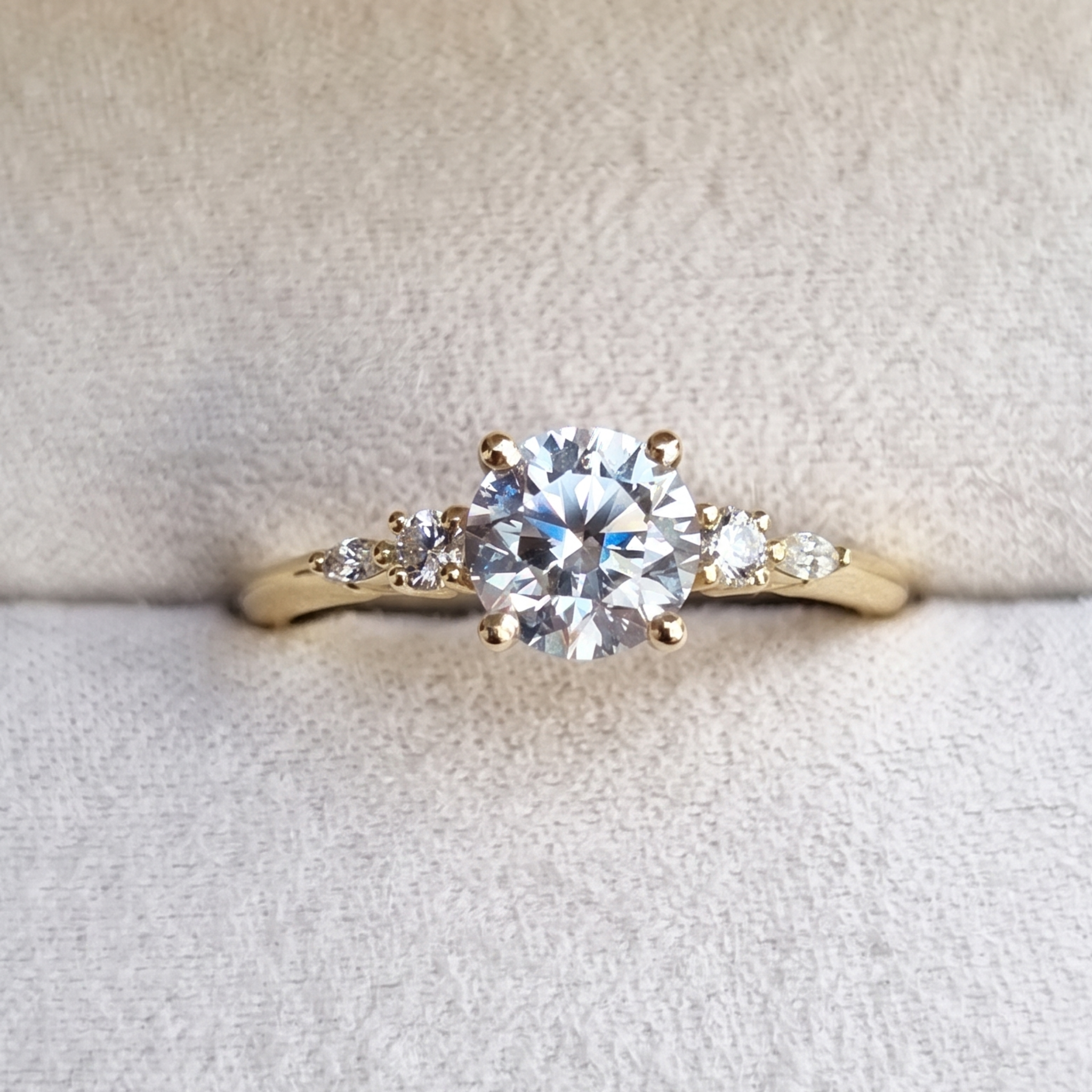A yellow gold solitaire ring with a round brilliant-cut center stone and small marquise-cut side stones, displayed on a textured cream-colored ring box.
