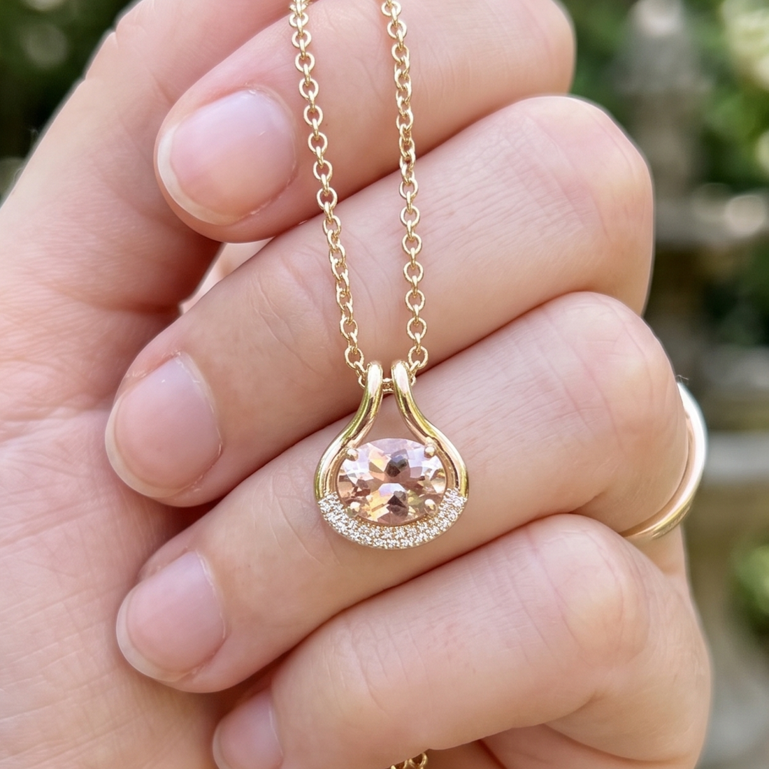 A gold teardrop-shaped pendant necklace with a central peach-colored gemstone and a halo of small clear stones, held in a person's hand.