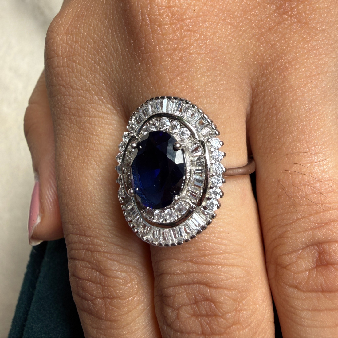 A close-up of a hand wearing a large oval ring with a deep blue center stone and surrounding silver-toned halo details.