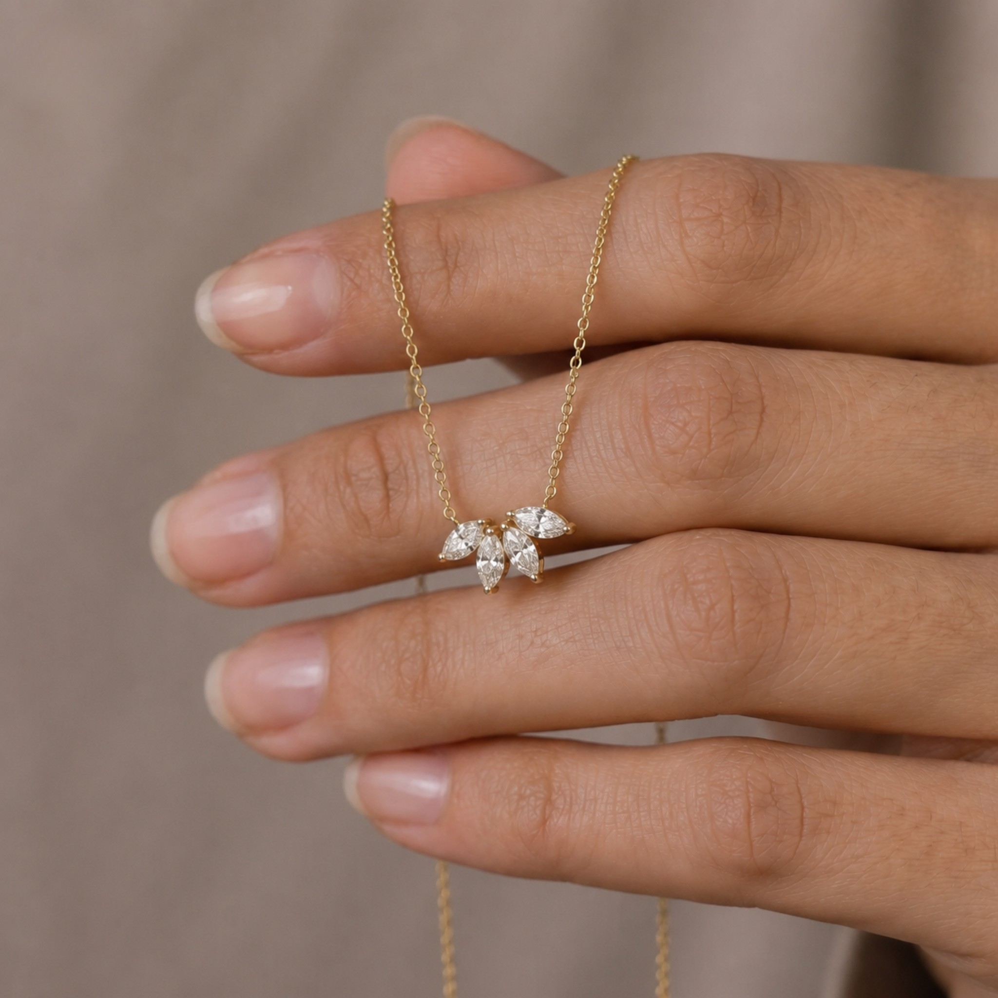 A delicate gold chain necklace with a small flower-shaped pendant set with clear stones, held on a person's fingers against a soft beige background.