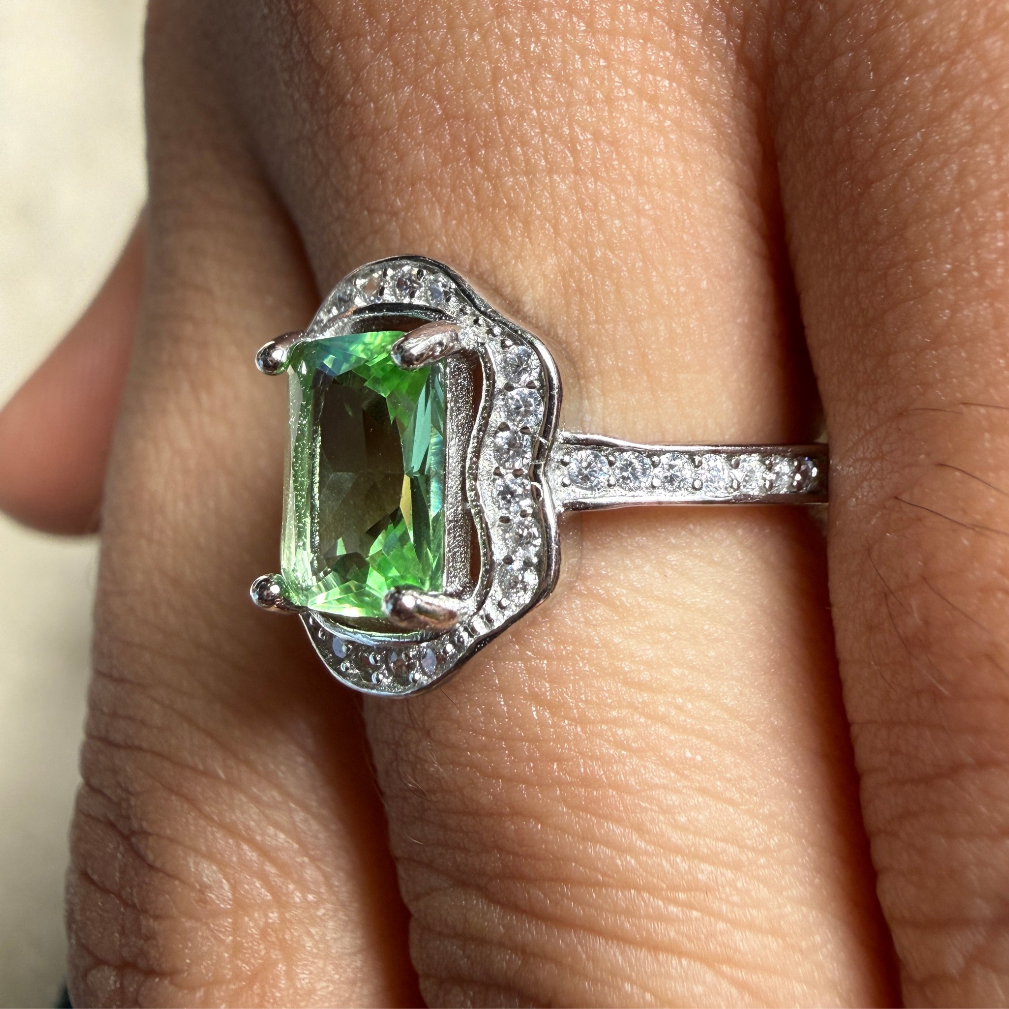 A close-up of a silver ring with a rectangular green gemstone and small clear accent stones, worn on a finger.