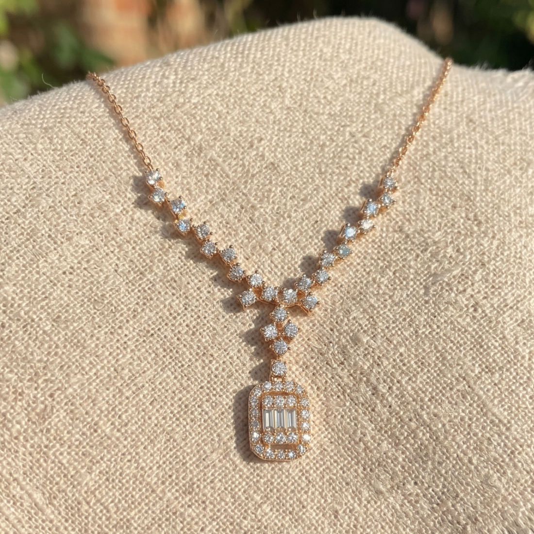A rose gold necklace with clear baguette and round-cut stones, displayed on a textured beige fabric stand.
