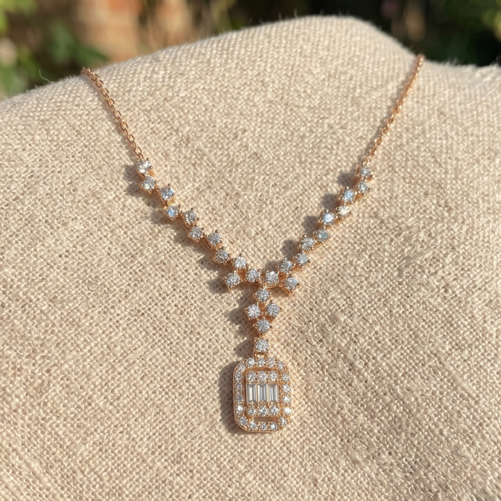 A rose gold necklace with clear baguette and round-cut stones, displayed on a textured beige fabric stand.