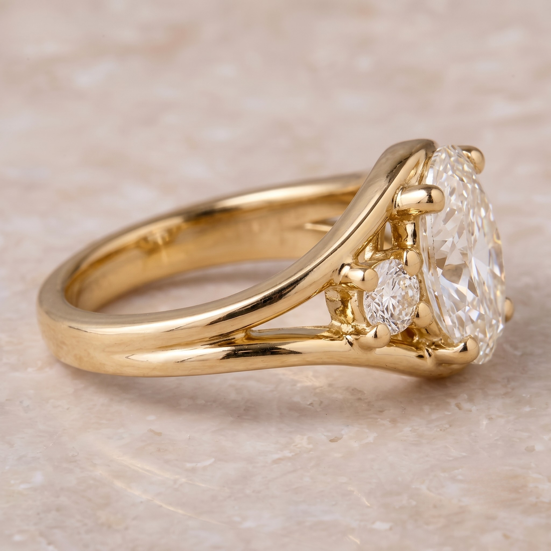A yellow gold ring featuring a large oval-cut clear gemstone in a prong setting, with a smaller round clear gemstone on the side, photographed on a light beige stone surface.