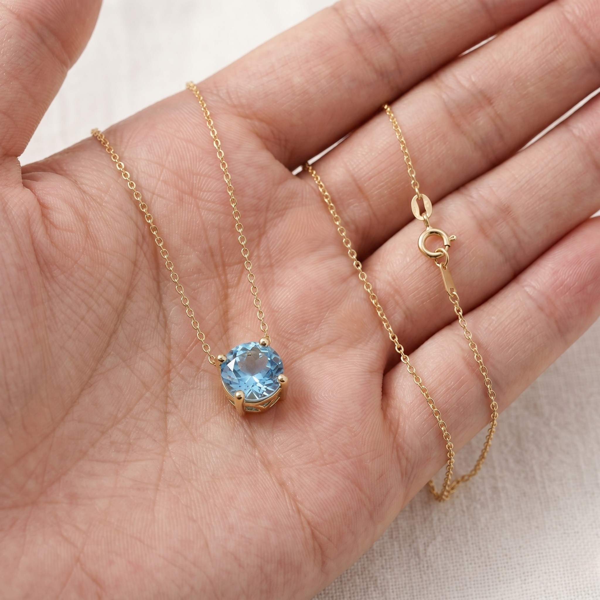 A delicate gold chain necklace with a round-cut light blue gemstone pendant, displayed on an open hand against a light neutral background.