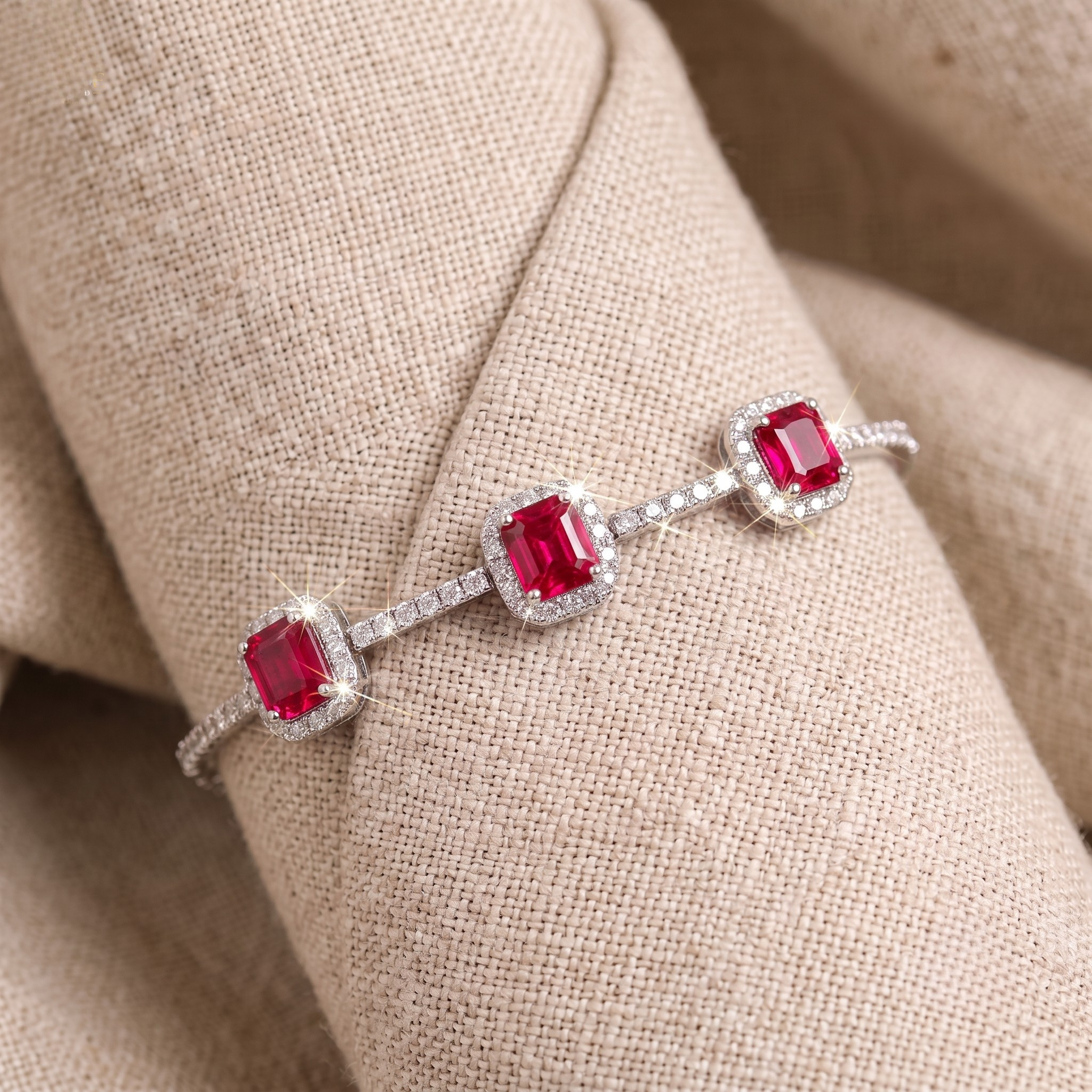 A silver-toned bracelet with three cushion-cut red gemstones surrounded by small clear stones, displayed on a beige fabric surface.
