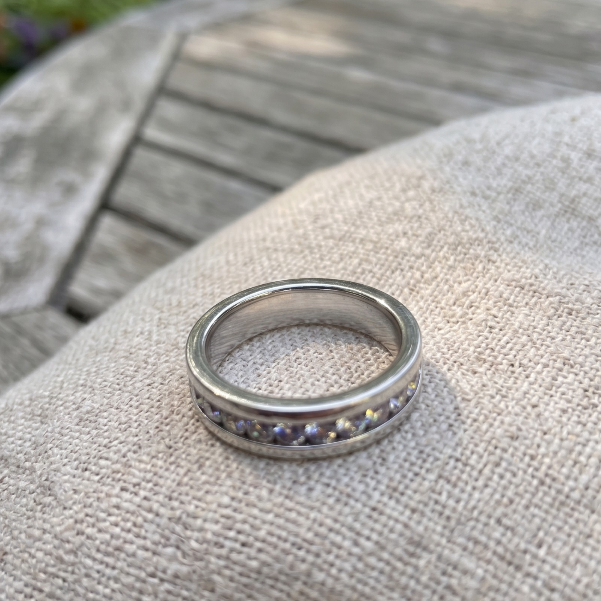 A silver band ring with a row of small round purple gemstones, resting on a textured beige fabric surface with a blurred wooden background.