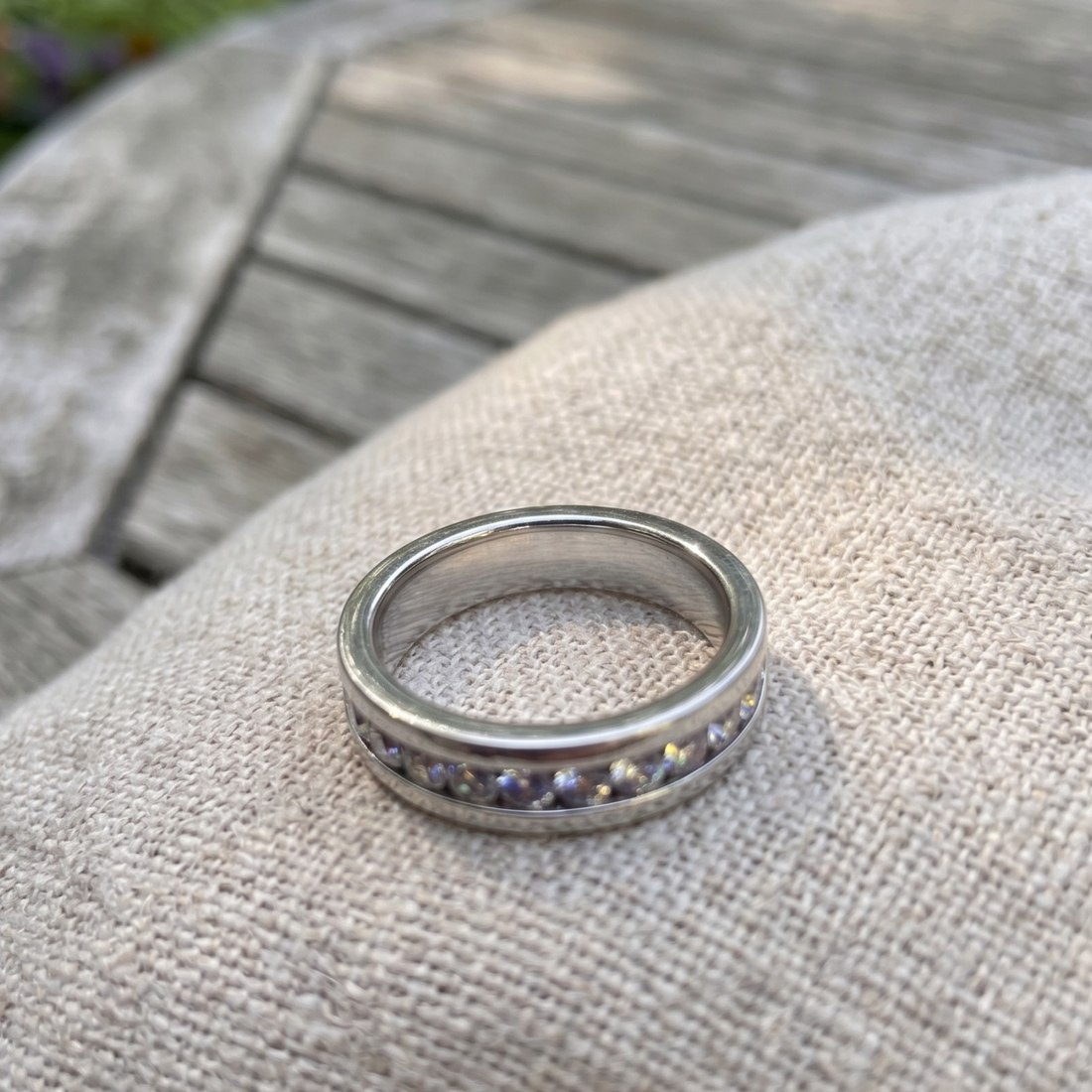 A silver band ring with a row of small round purple gemstones, resting on a textured beige fabric surface with a blurred wooden background.