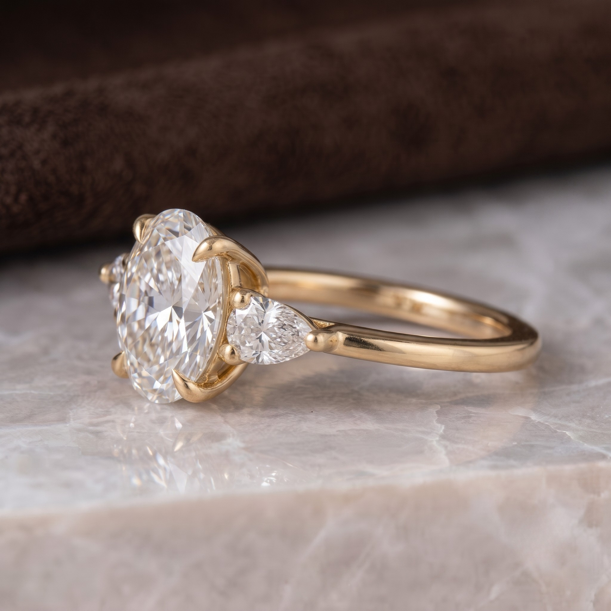 A yellow gold engagement ring with a large oval center stone and a smaller pear-cut side stone, resting on a light marble surface with a dark brown textured background.